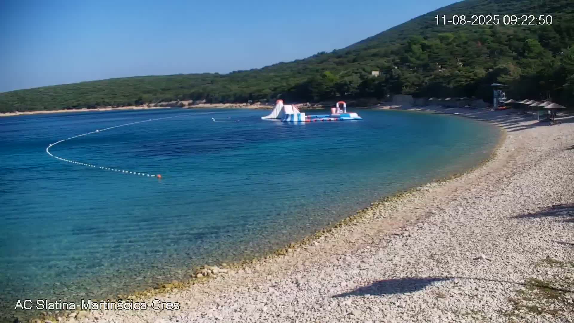 A pebble beach curves around clear, turquoise water with an inflatable water park visible offshore on a sunny day.