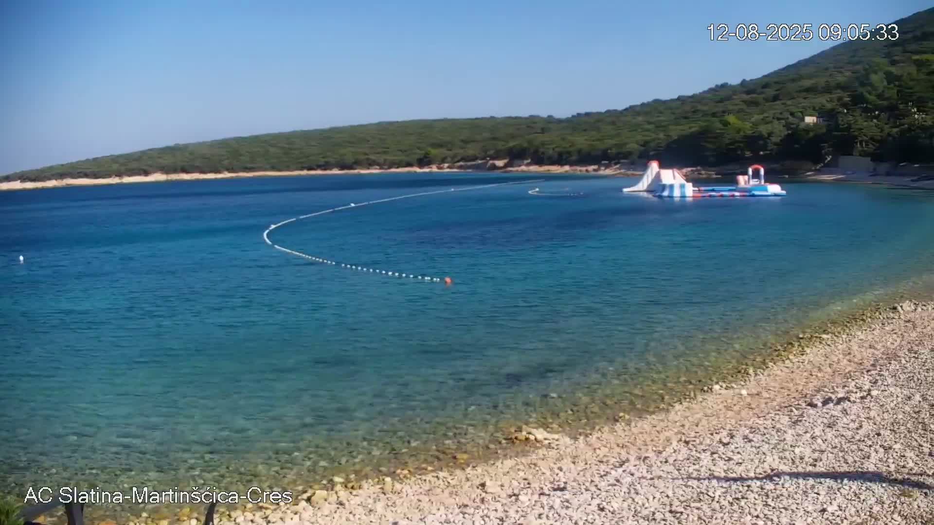 A pebble beach with clear, blue water and an inflatable water park is visible on a sunny day.