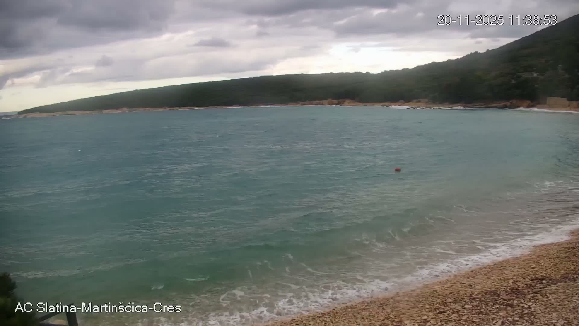 A pebble beach lines a calm, turquoise bay with a red buoy, backed by a lush green forested hillside under a heavily overcast sky.