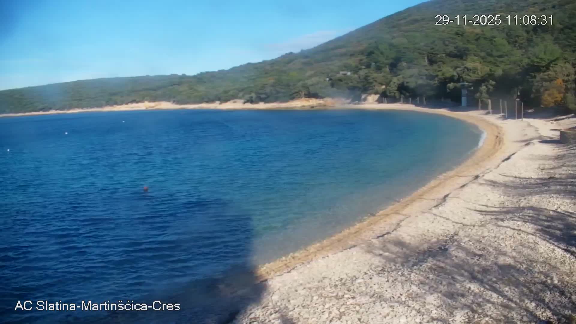 A sunny day illuminates a tranquil blue bay with a curving pebble beach, backed by a lush green forested hillside under a clear blue sky.