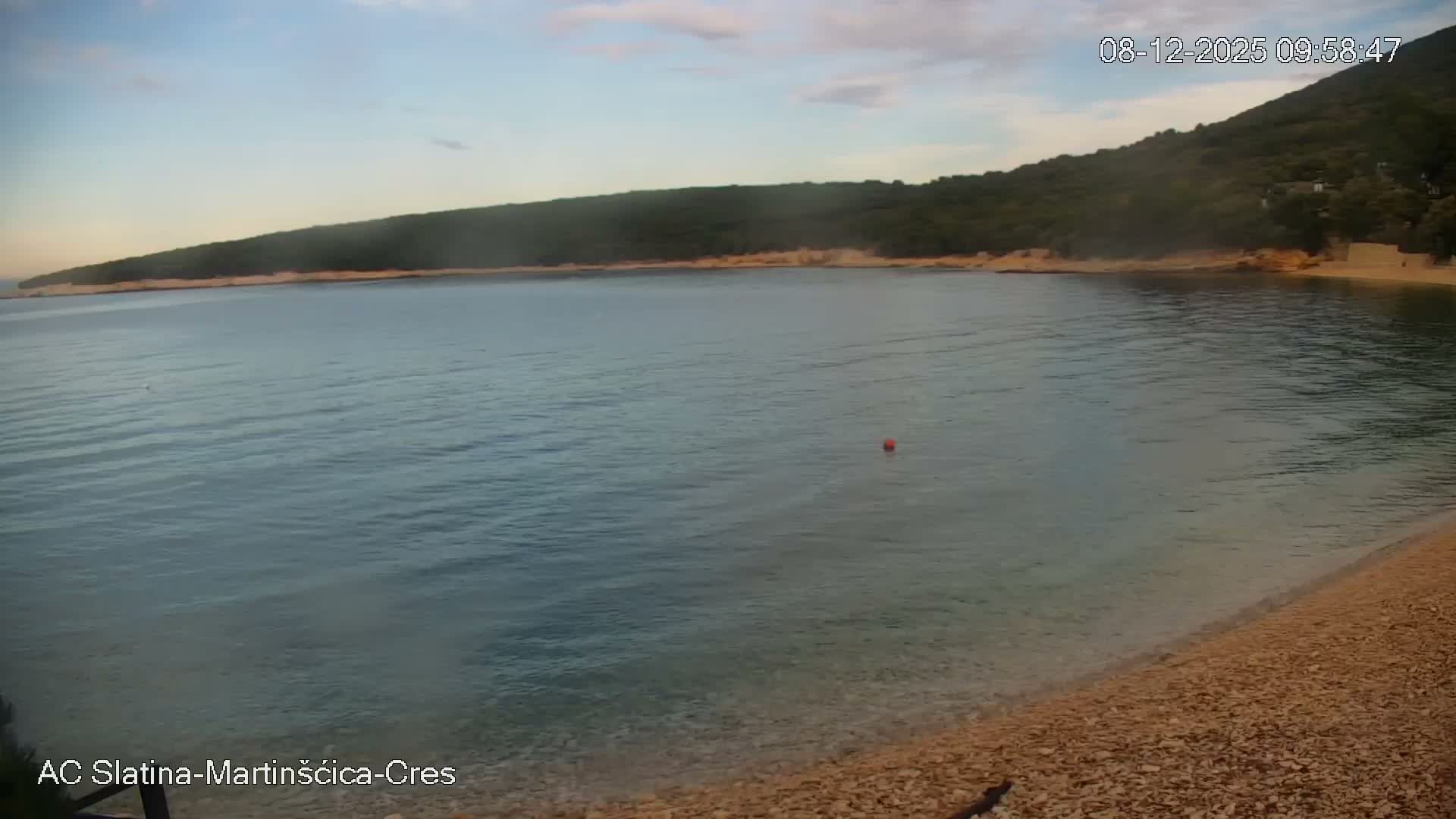 A calm, clear bay with a pebble beach in the foreground and a tree-covered hillside across the water is visible under a partly cloudy sky, with a single red buoy floating in the tranquil water.