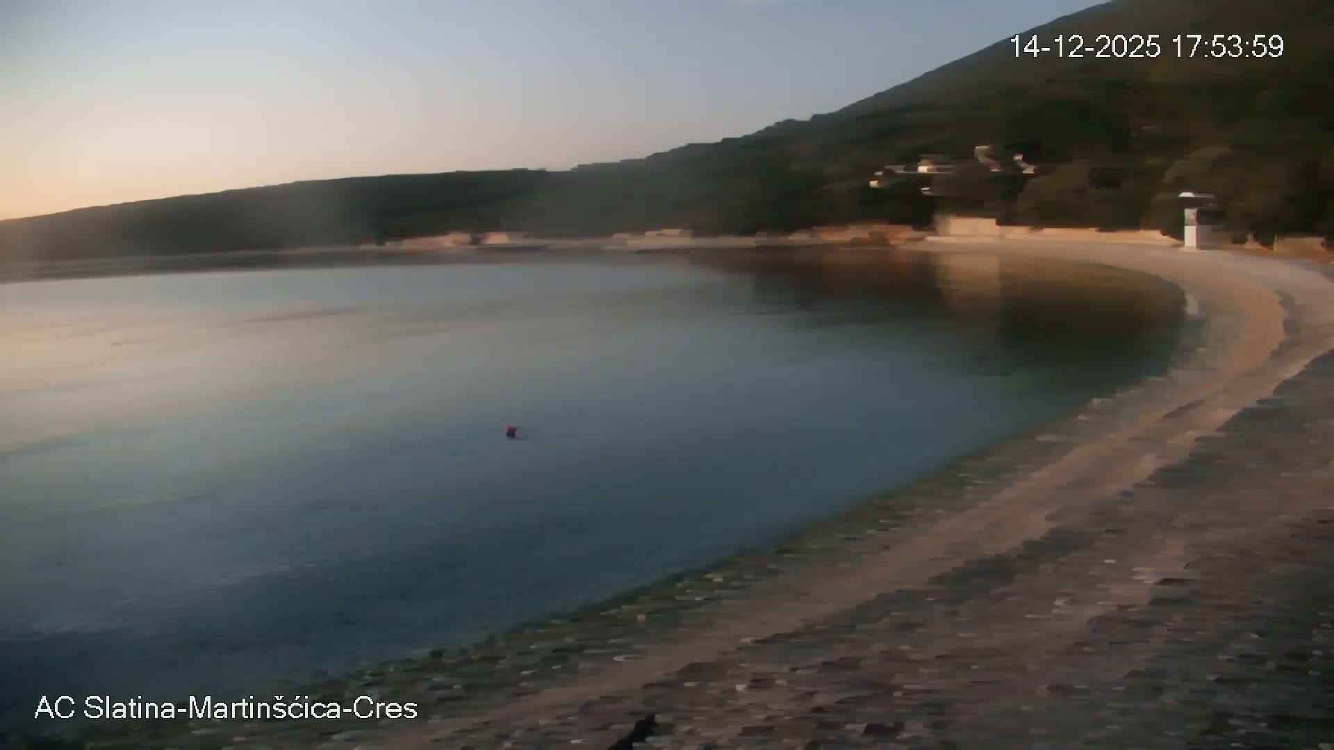 A calm, clear bay with a pebble beach in the foreground and a tree-covered hillside across the water is visible under a partly cloudy sky, with a single red buoy floating in the tranquil water.