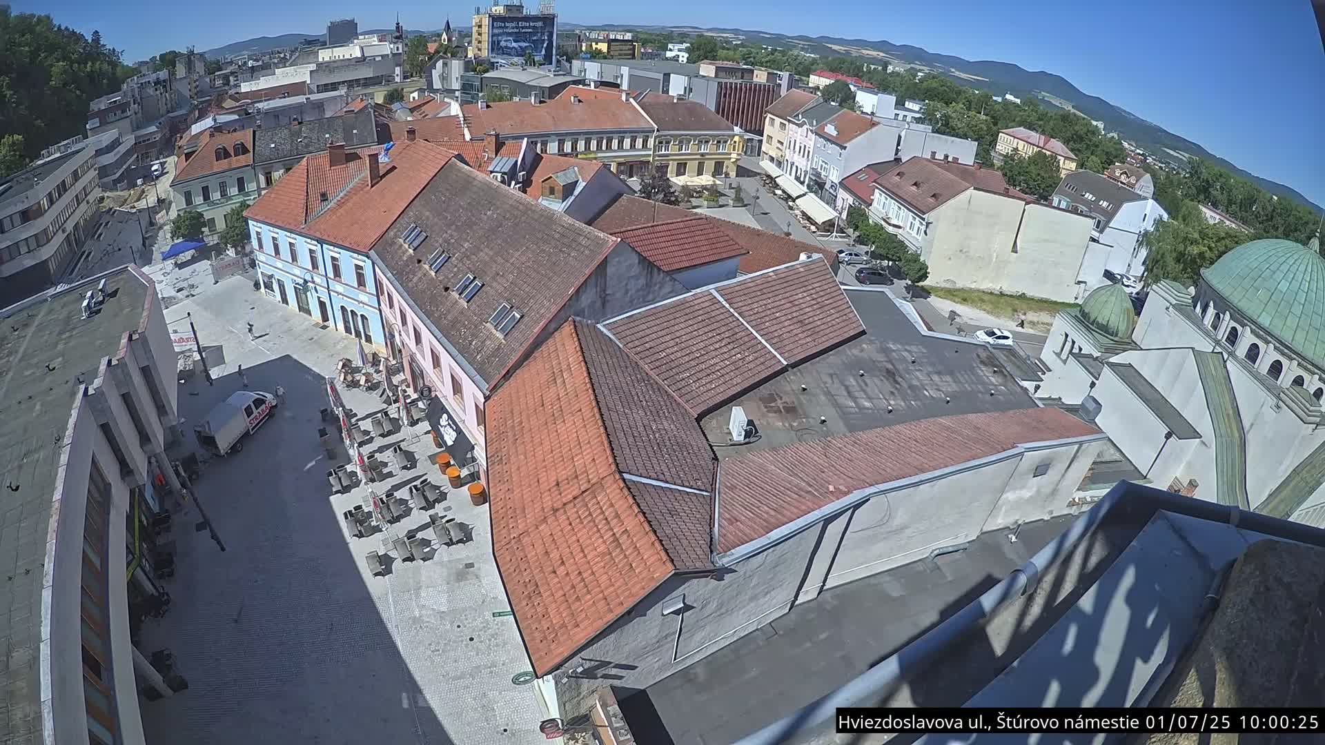 A high-angle, sunny view of a town square with several buildings, outdoor seating, and a vehicle, all surrounded by more buildings and trees.