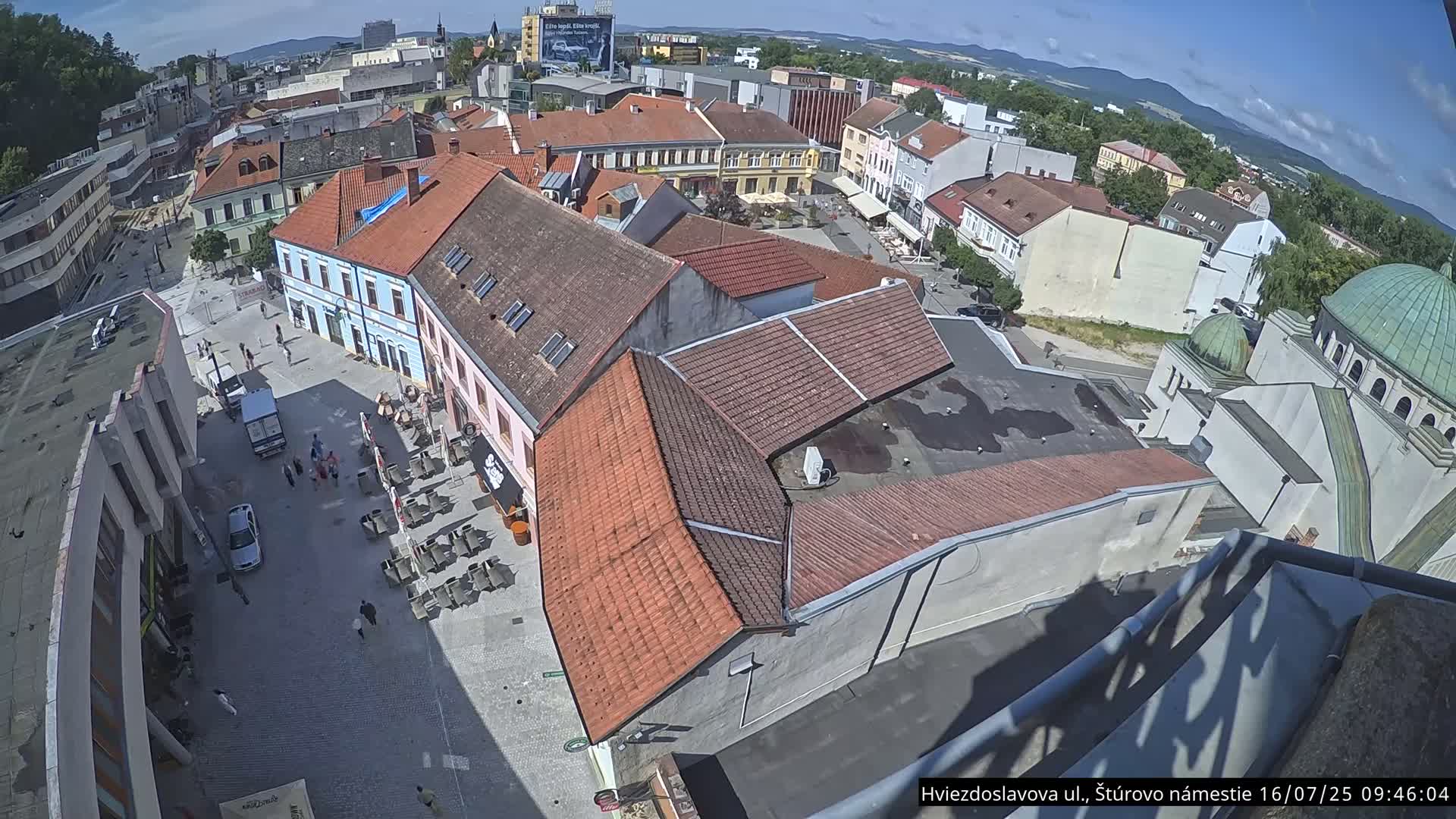An aerial view of a town square on a sunny day shows several buildings, a few people walking, and outdoor seating at cafes.