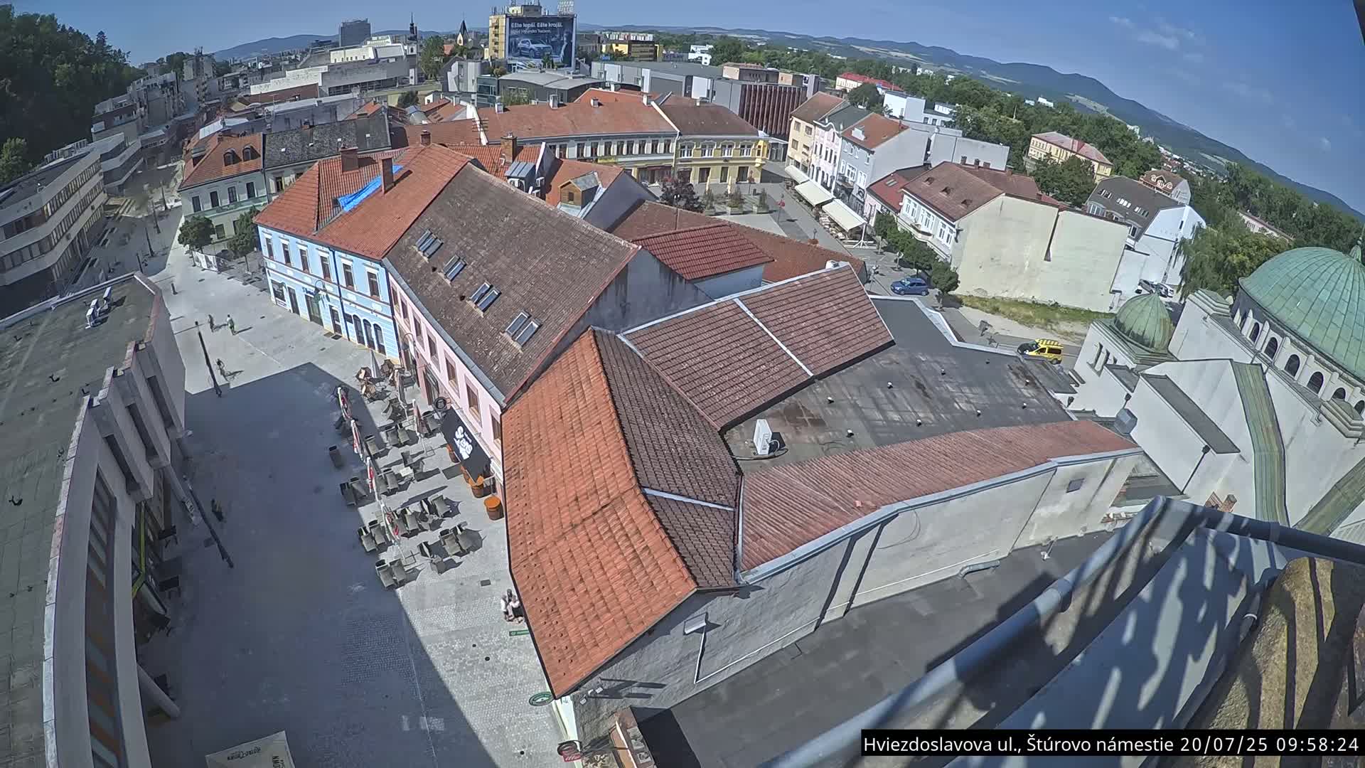 A high-angle view of a town square with numerous buildings under a clear, sunny sky, featuring outdoor seating at a café and a partially visible dome-shaped structure.