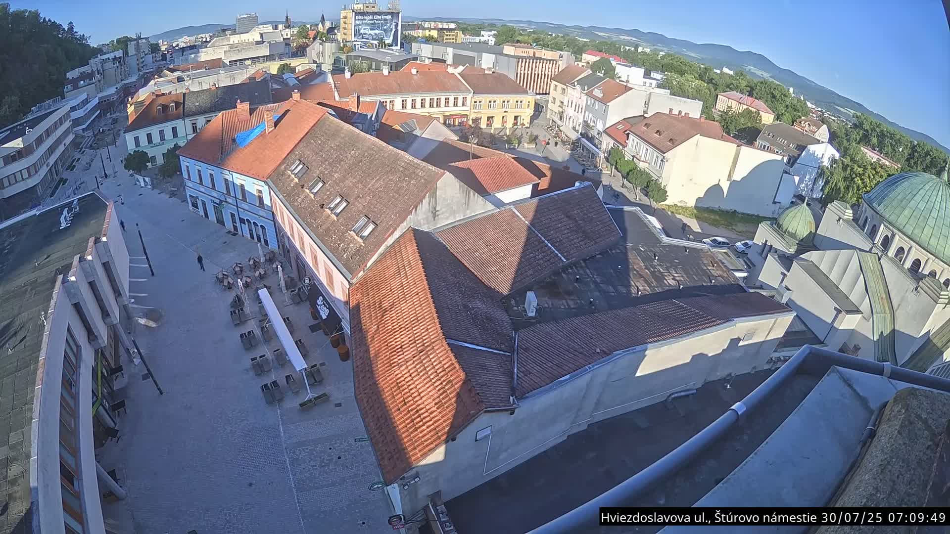 A high-angle view of a town square with numerous buildings under a clear, sunny sky, featuring outdoor seating at a café and a partially visible dome-shaped structure.