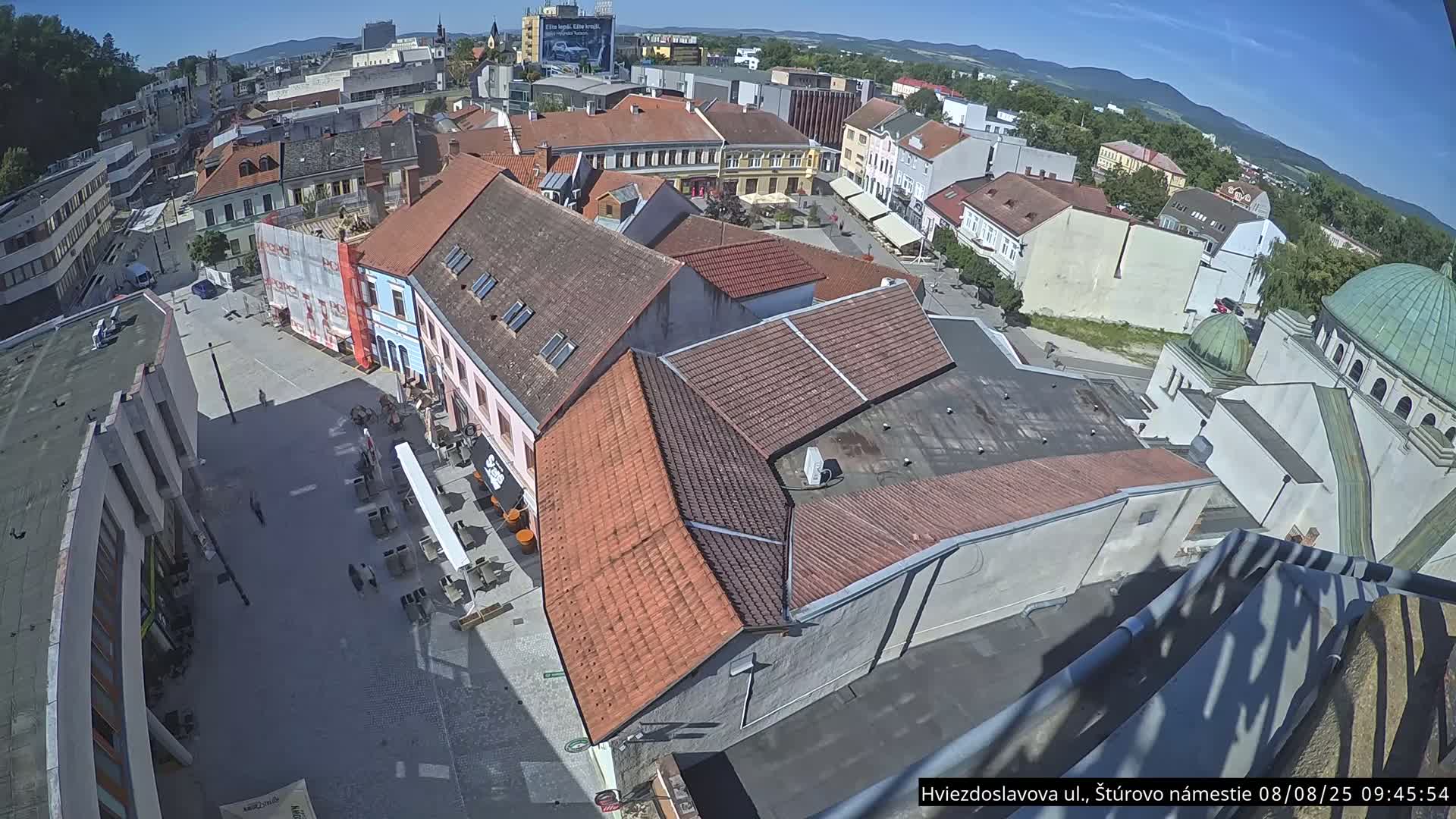 A high-angle, sunny view of a town square with several buildings, including a building with a dome-shaped roof, under a clear blue sky.