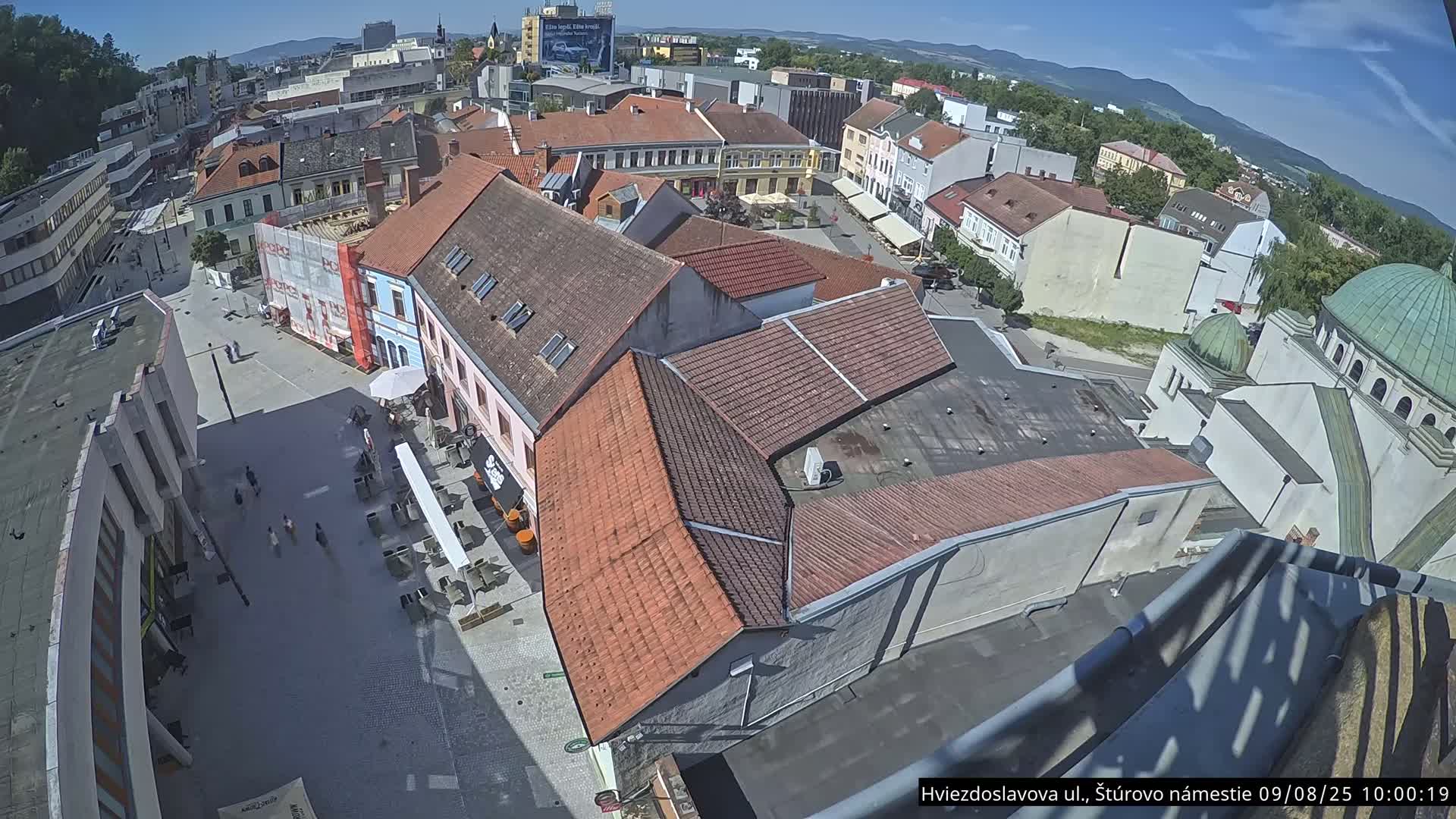 A high-angle, sunny view of a town square surrounded by buildings with red tile roofs, one of which is under construction, and a partially visible dome-shaped structure.