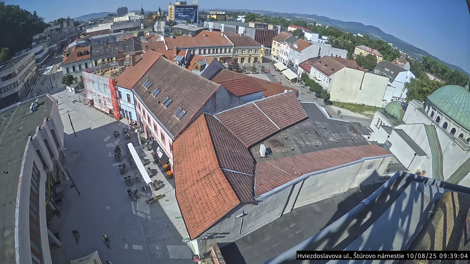 An elevated view shows a town square with buildings, some under construction, on a sunny day.