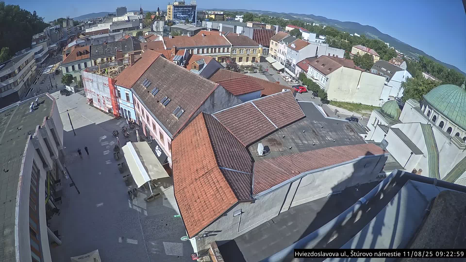 A high-angle, sunny view shows a town square with red-tiled buildings, a few people, and a partially visible synagogue in the background.