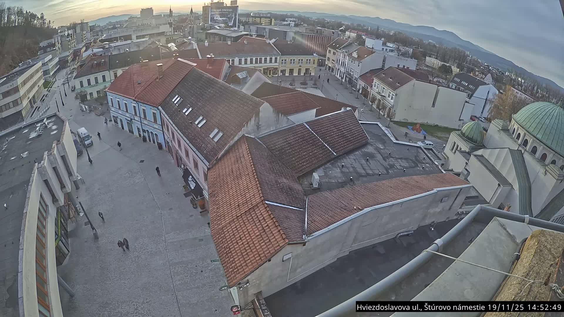 A high-angle, sunny view shows a town square with red-tiled buildings, a few people, and a partially visible synagogue in the background.