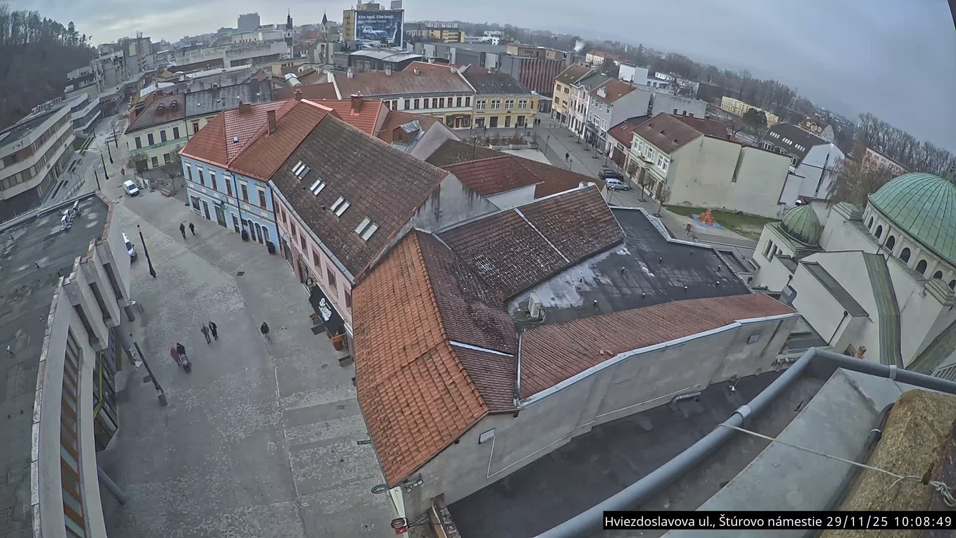 An overcast and cloudy day casts a soft light over an elevated view of a bustling European town square featuring traditional buildings with tiled roofs, a green-domed structure, and numerous pedestrians strolling along the paved streets.