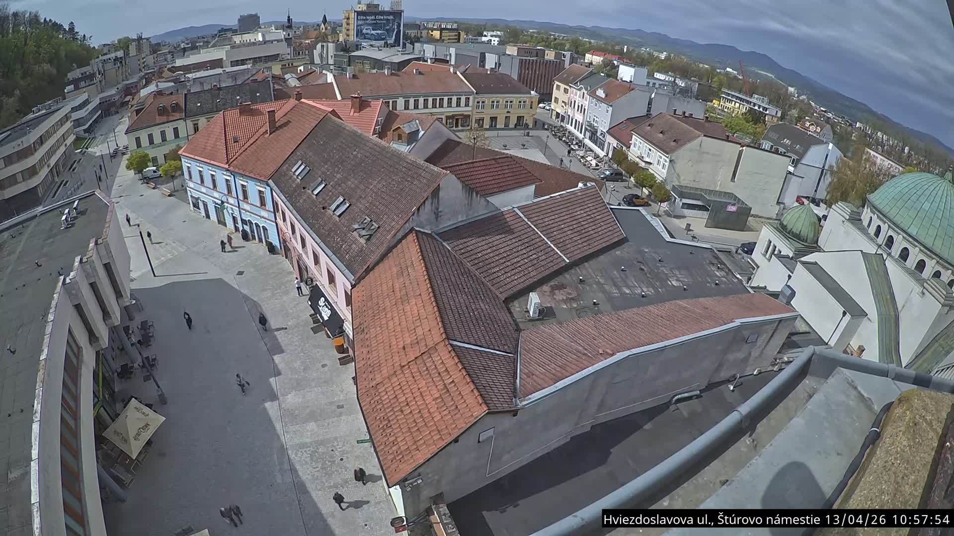 An overcast and cloudy day casts a soft light over an elevated view of a bustling European town square featuring traditional buildings with tiled roofs, a green-domed structure, and numerous pedestrians strolling along the paved streets.