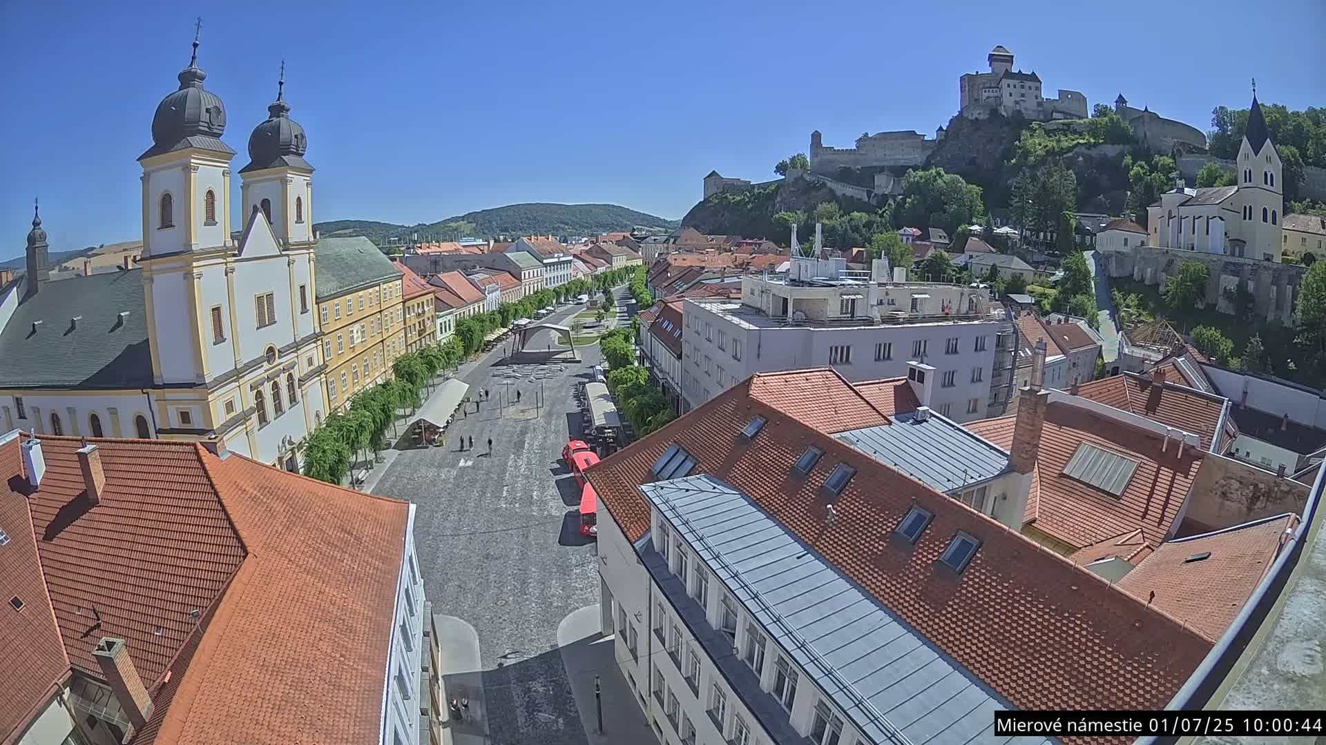 A high-angle view of a town square on a sunny day, with a large church and a castle visible on a hill in the background.