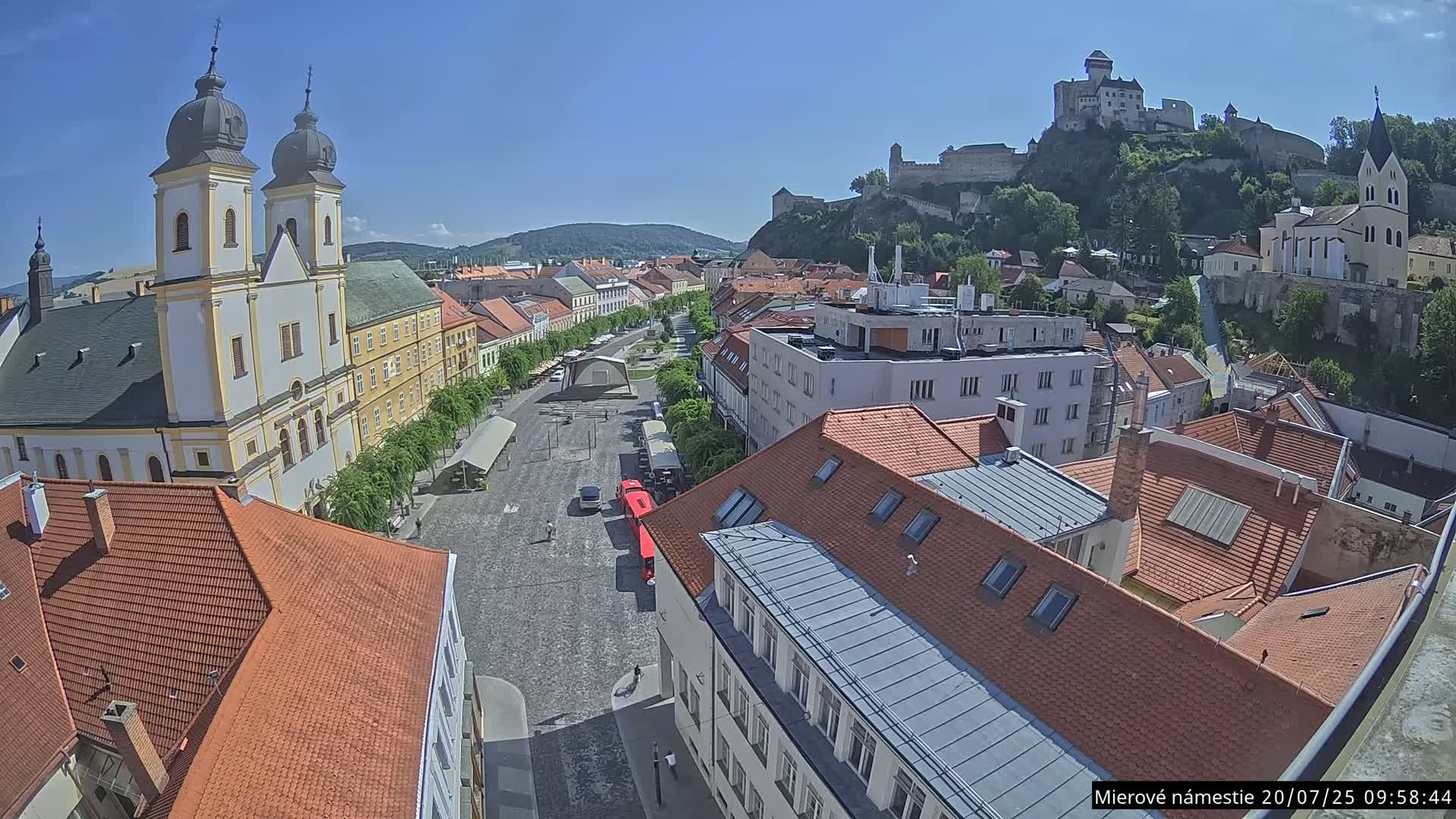 A high-angle view on a sunny day shows a town square with a church, buildings, and a castle on a hill in the background.