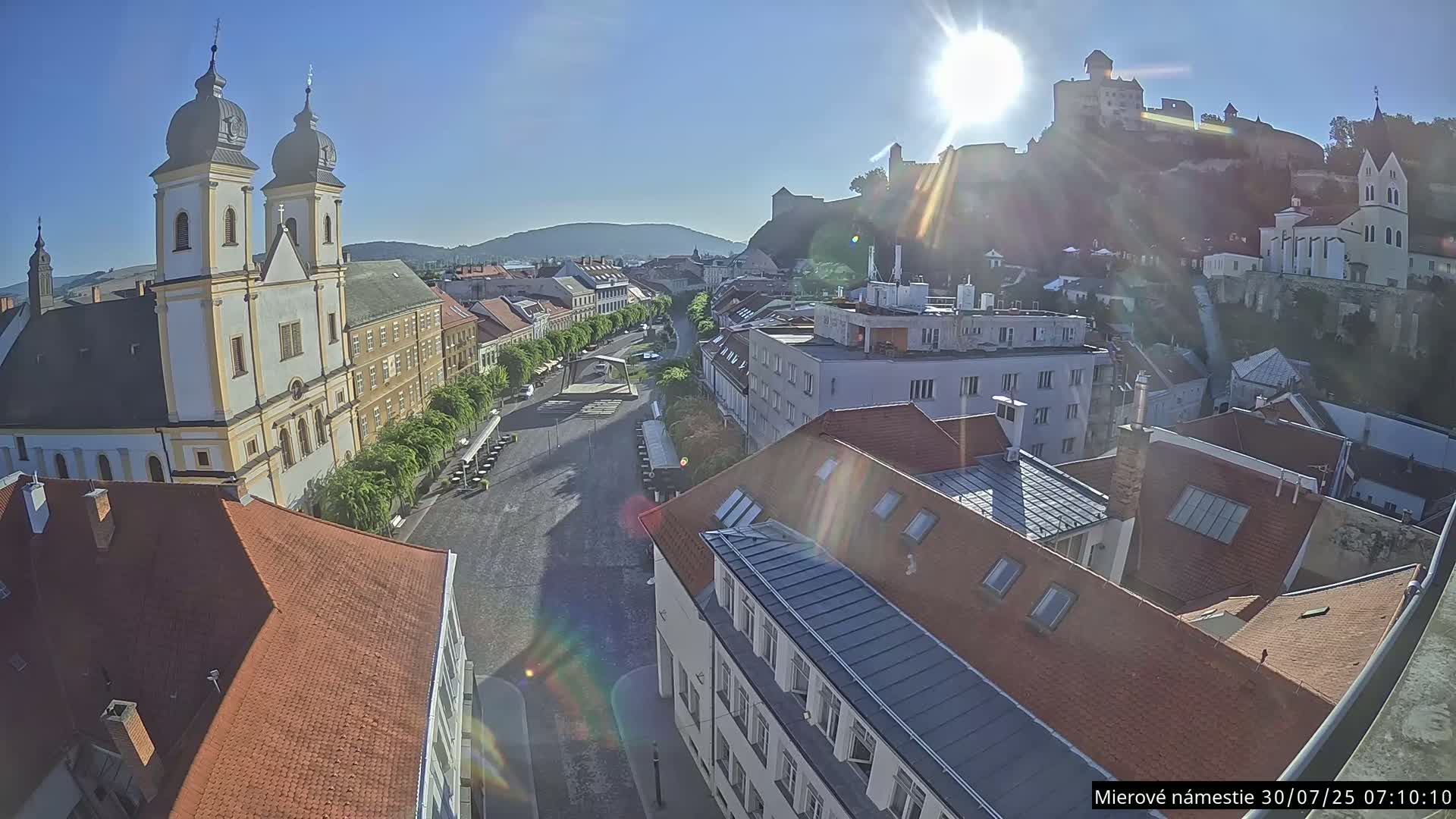 A high-angle view on a sunny day shows a town square with a church, buildings, and a castle on a hill in the background.