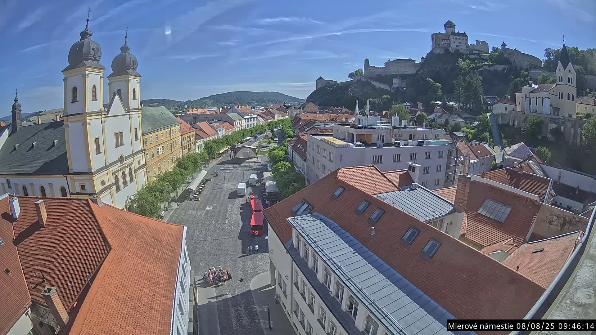 An elevated view shows a town square with a church, buildings, a red vehicle, and a castle on a hill in the background under sunny skies.