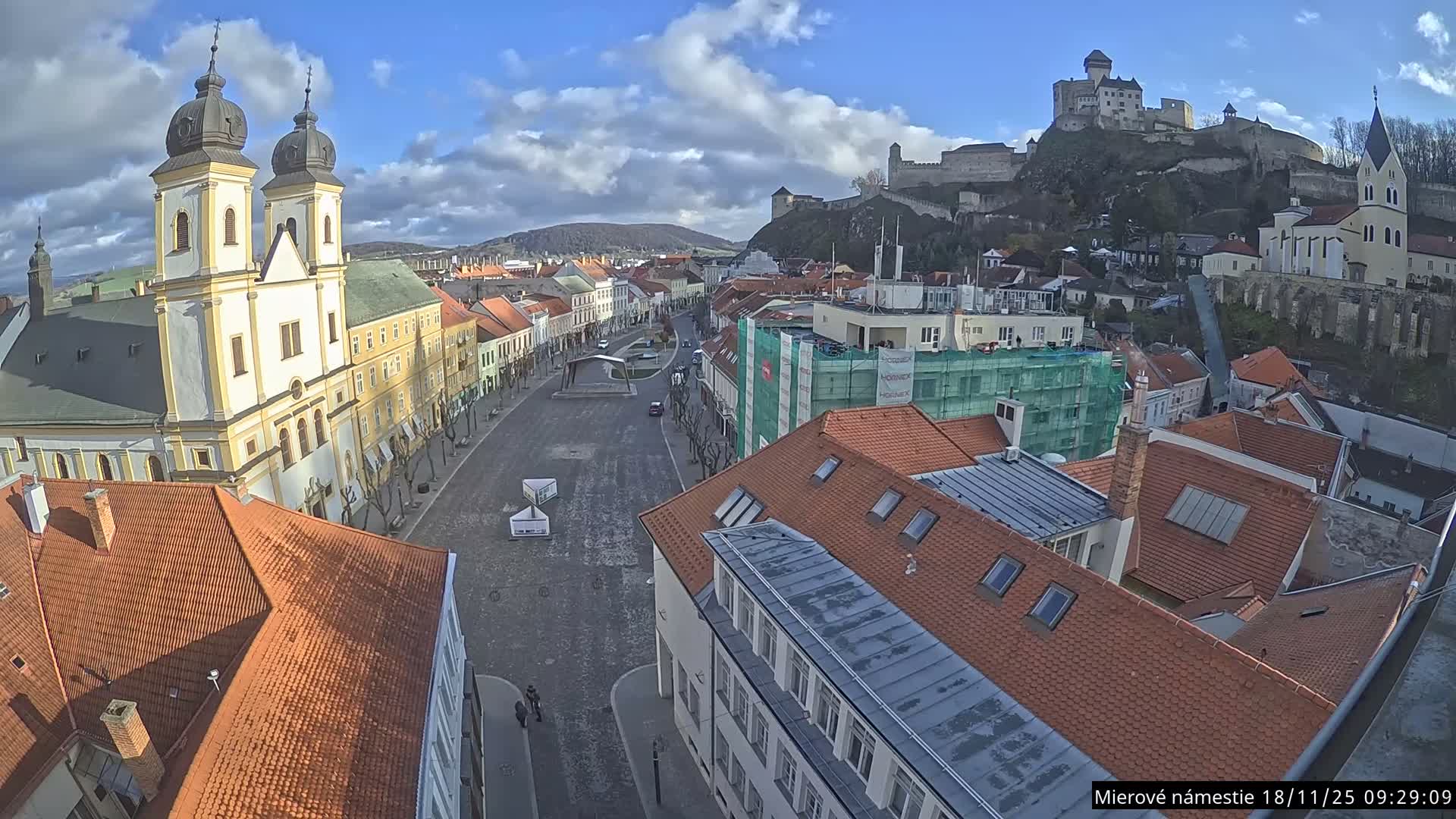 An elevated view reveals a historic European townscape, featuring a prominent twin-towered church on the left, a wide central street lined with colorful buildings, and a large castle dominating a distant hill to the right, all under a partly cloudy sky.