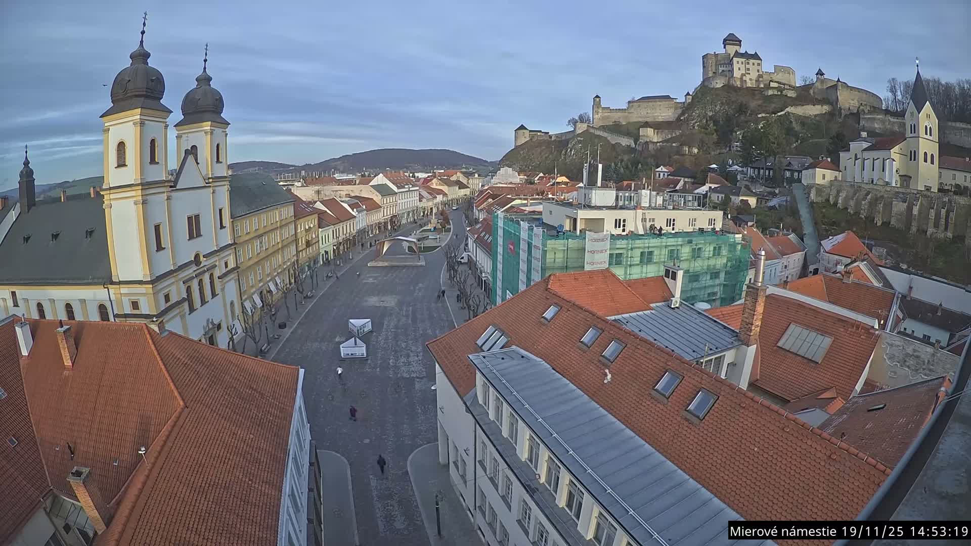 An elevated view reveals a historic European townscape, featuring a prominent twin-towered church on the left, a wide central street lined with colorful buildings, and a large castle dominating a distant hill to the right, all under a partly cloudy sky.