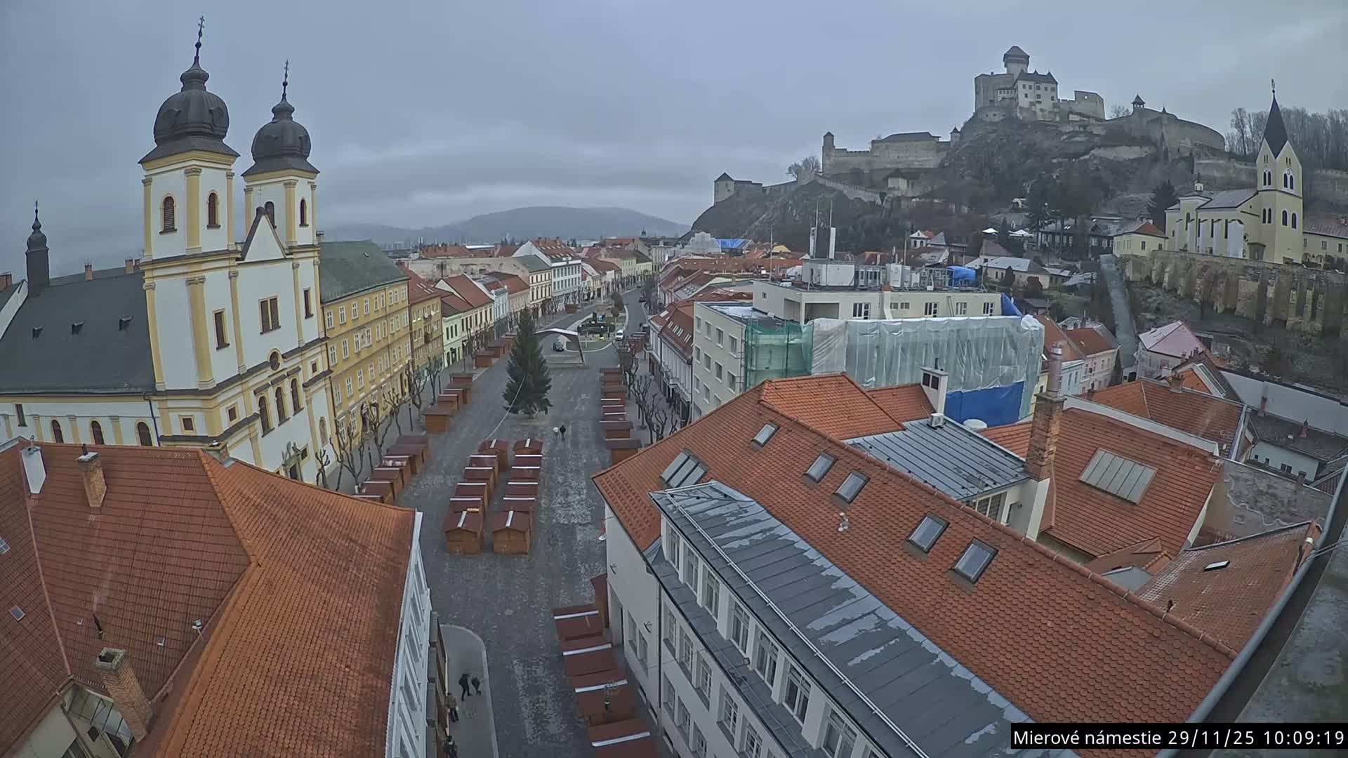 A high-angle view reveals a bustling historic European town square on a grey, overcast day, featuring a grand church with two dark-domed towers on the left, a central Christmas tree surrounded by numerous market stalls, and a prominent castle perched atop a distant hill.