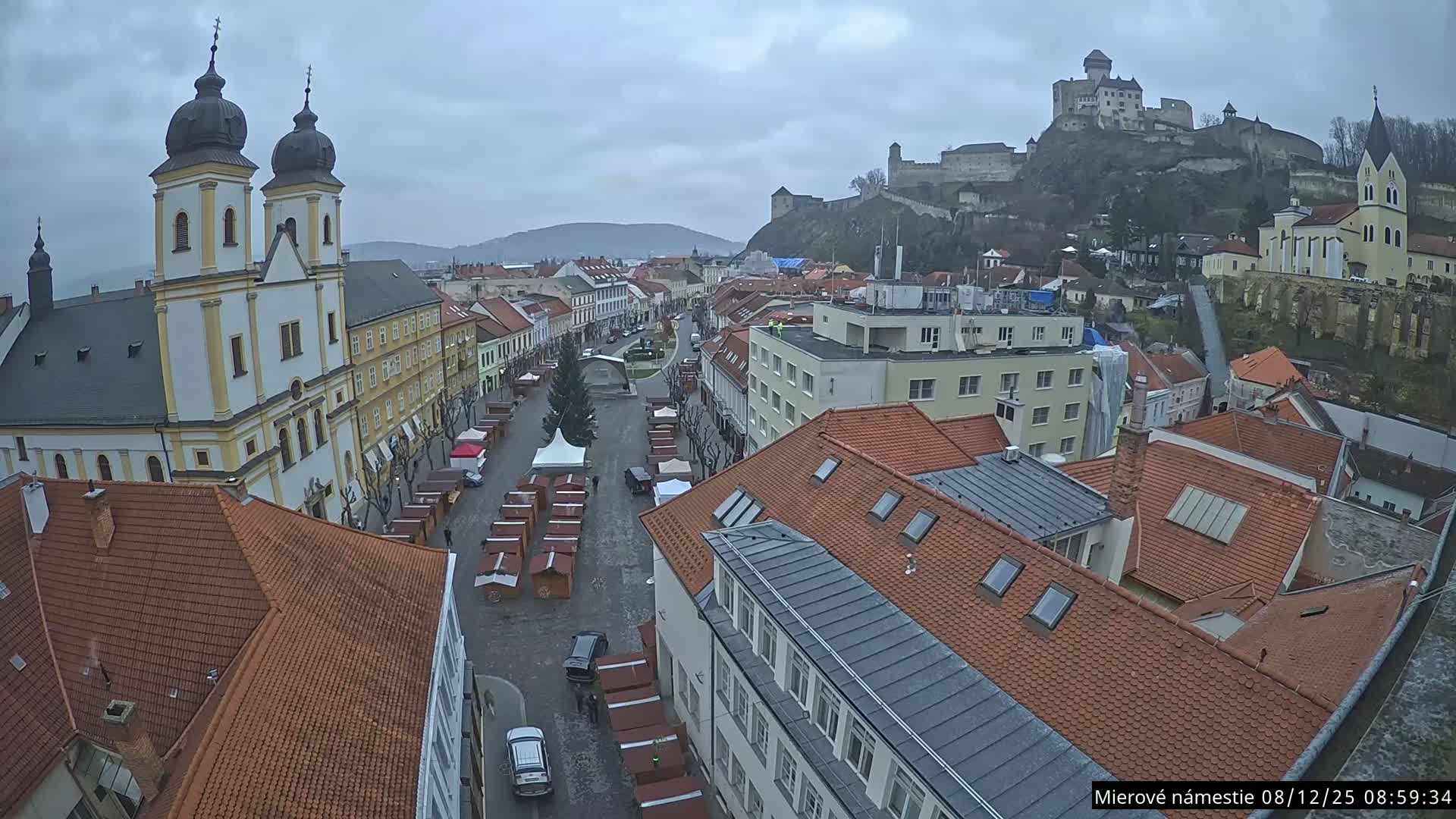 An elevated wide shot captures a European town square lined with market stalls and a Christmas tree, featuring a large church with twin domed spires on the left and a formidable castle on a hill in the distance, all under an overcast sky.