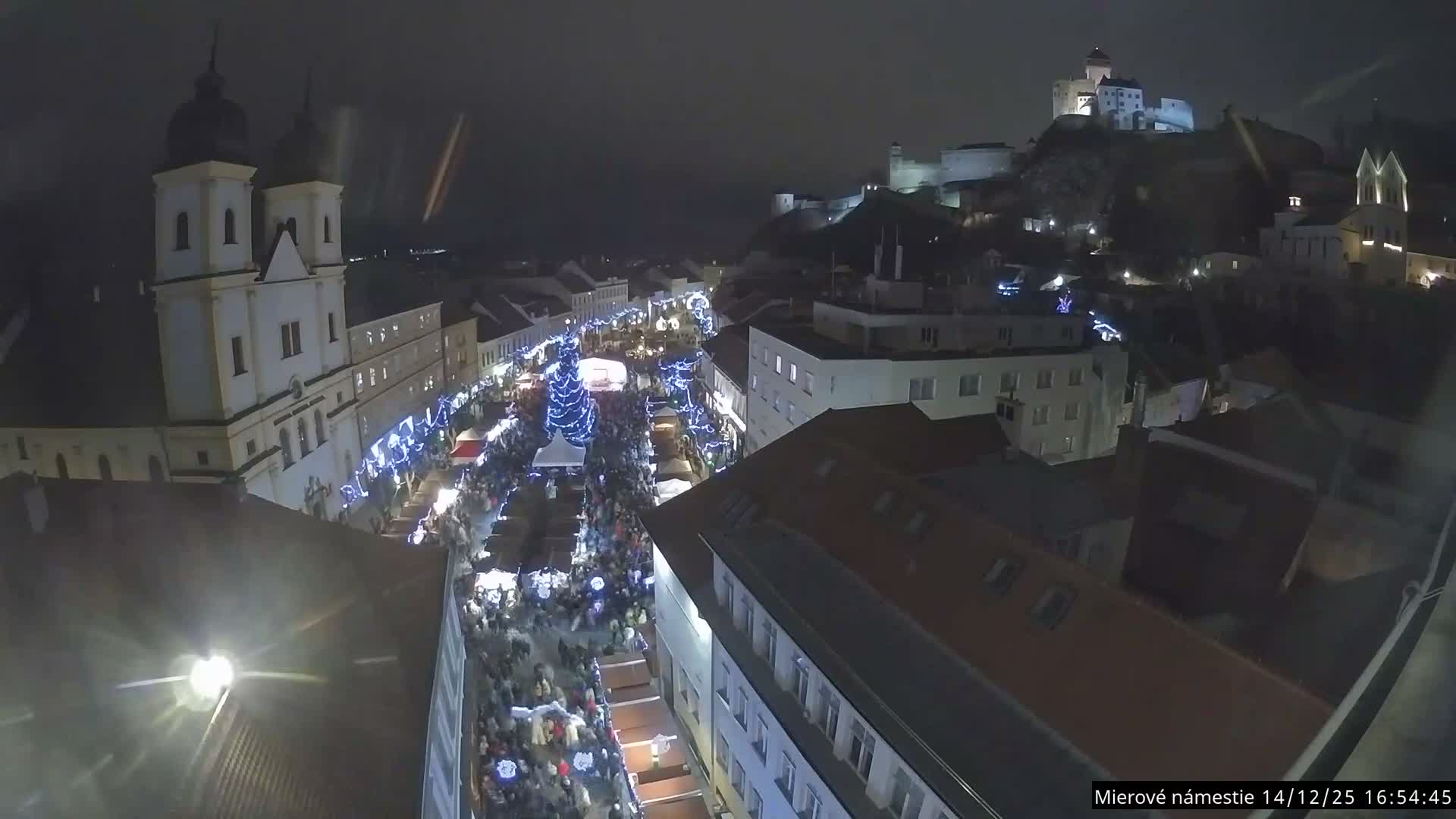 An elevated wide shot captures a European town square lined with market stalls and a Christmas tree, featuring a large church with twin domed spires on the left and a formidable castle on a hill in the distance, all under an overcast sky.