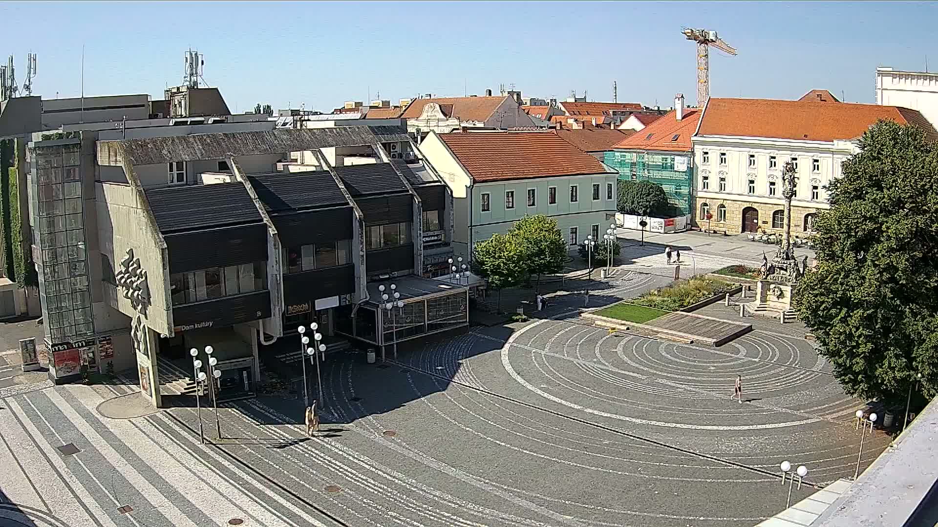 A sunlit town square with a circular, patterned pavement features buildings surrounding it, including a multi-story structure with dark roofing, and a construction crane is visible in the background.
