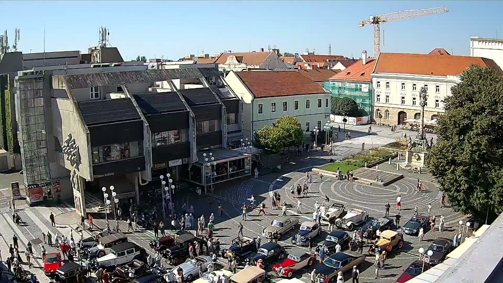 A town square on a sunny day filled with vintage cars and people, surrounded by buildings including a multi-story building with a dark facade and a taller building with a red roof.