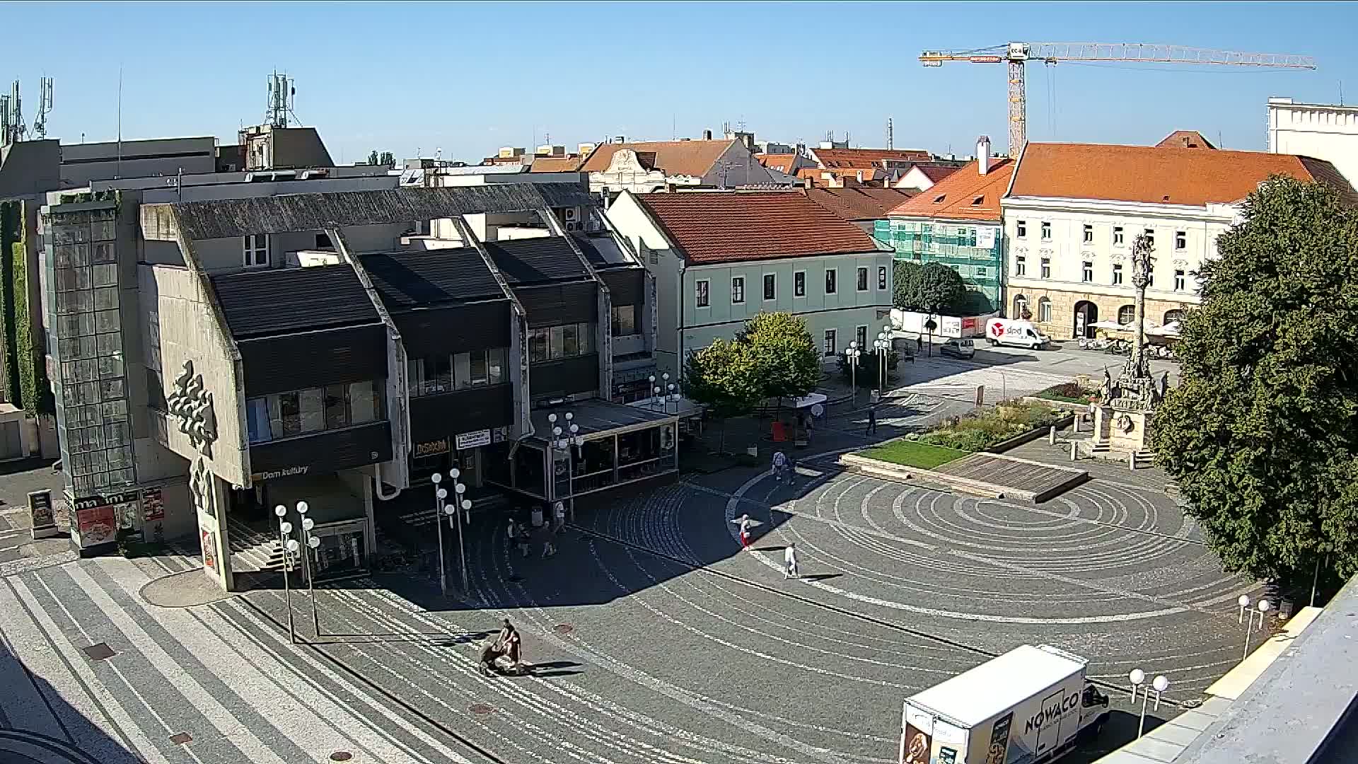 A sunny day reveals a town square with a circular, patterned pavement, surrounded by buildings, a monument, and a few pedestrians.