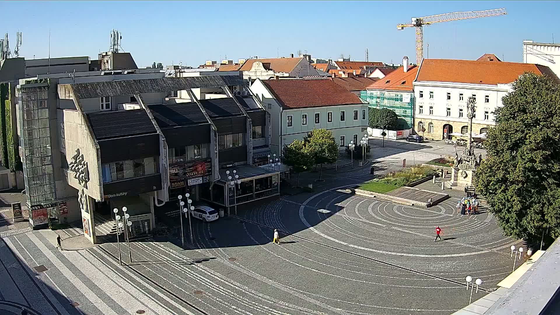 A high-angle view shows a town square on a sunny day with a circular paved area, buildings surrounding it, a monument in the center, and a construction crane in the background.