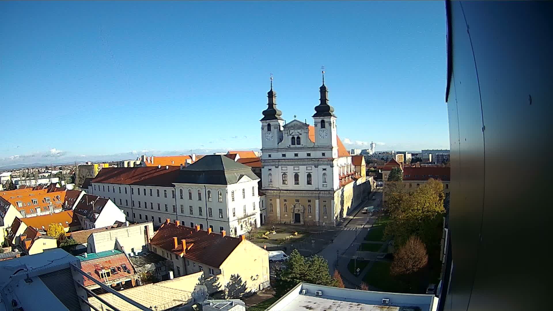 An aerial view of a European town features a prominent white church with twin dark-domed towers and clock faces, surrounded by buildings with reddish-brown roofs and some trees, all under a clear blue, sunny sky.