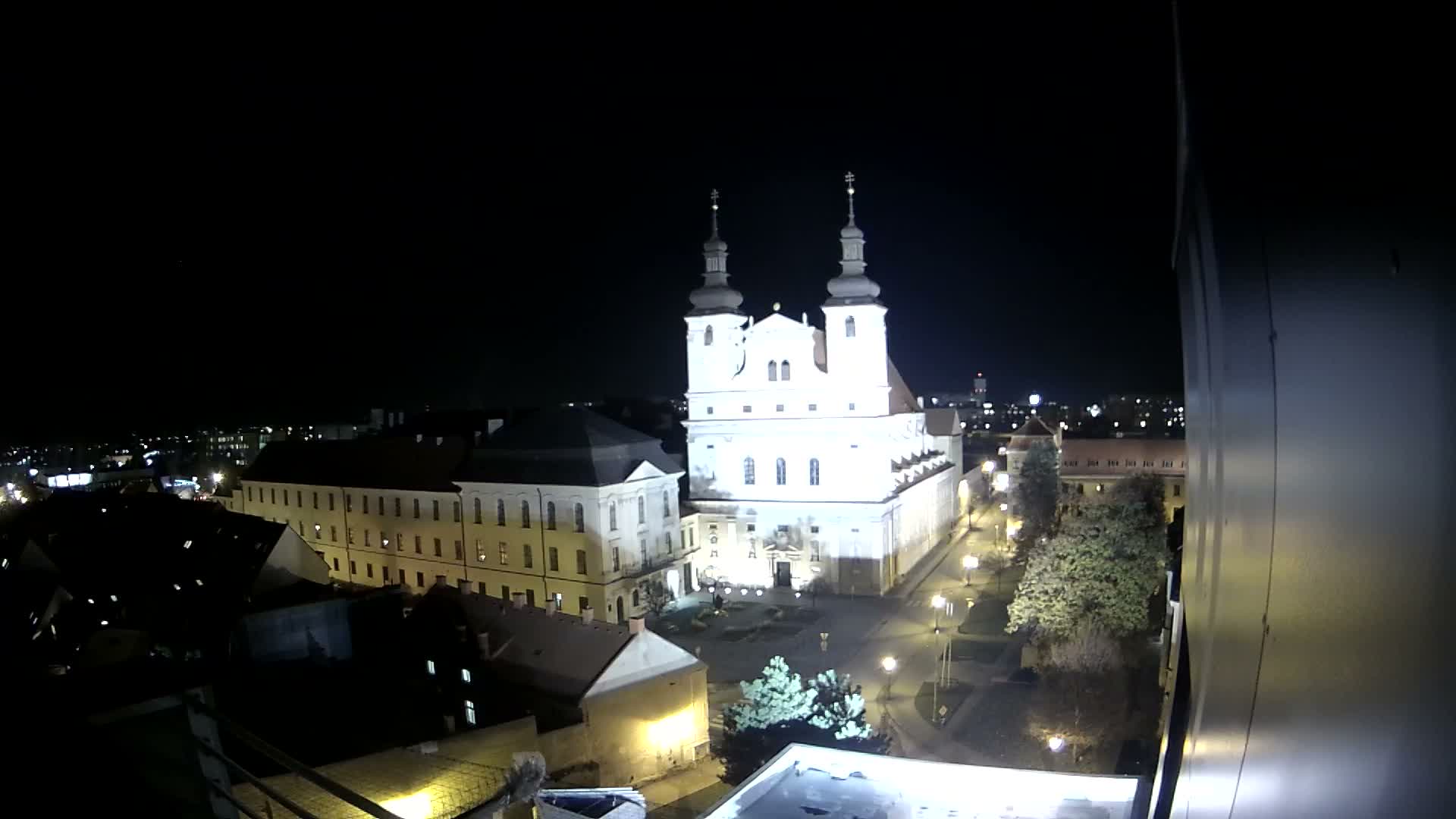 An aerial view of a European town features a prominent white church with twin dark-domed towers and clock faces, surrounded by buildings with reddish-brown roofs and some trees, all under a clear blue, sunny sky.