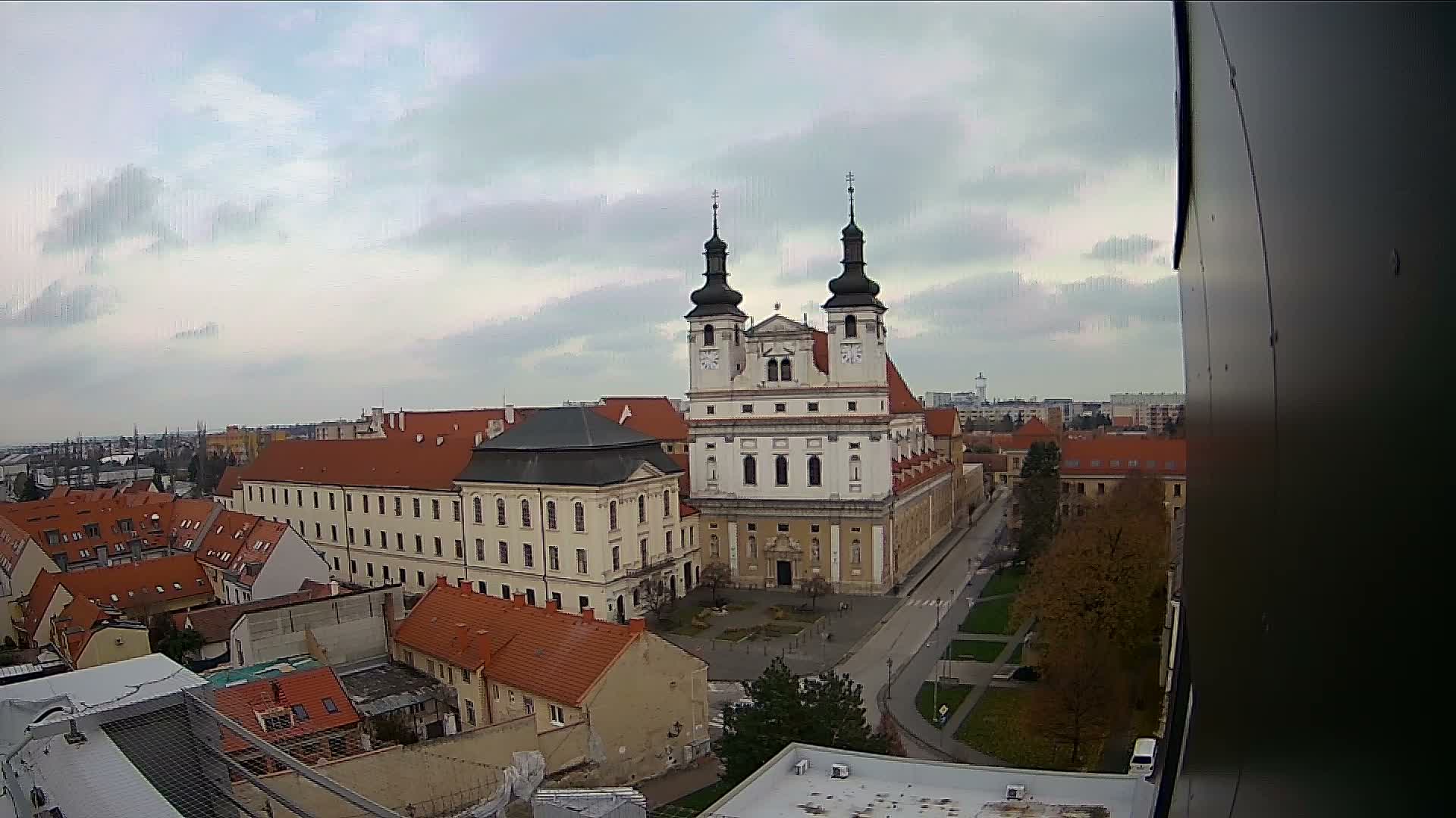 An elevated view reveals a European townscape centered around an ornate, two-spired church, surrounded by buildings with red tile roofs and autumn trees, all under an overcast sky.