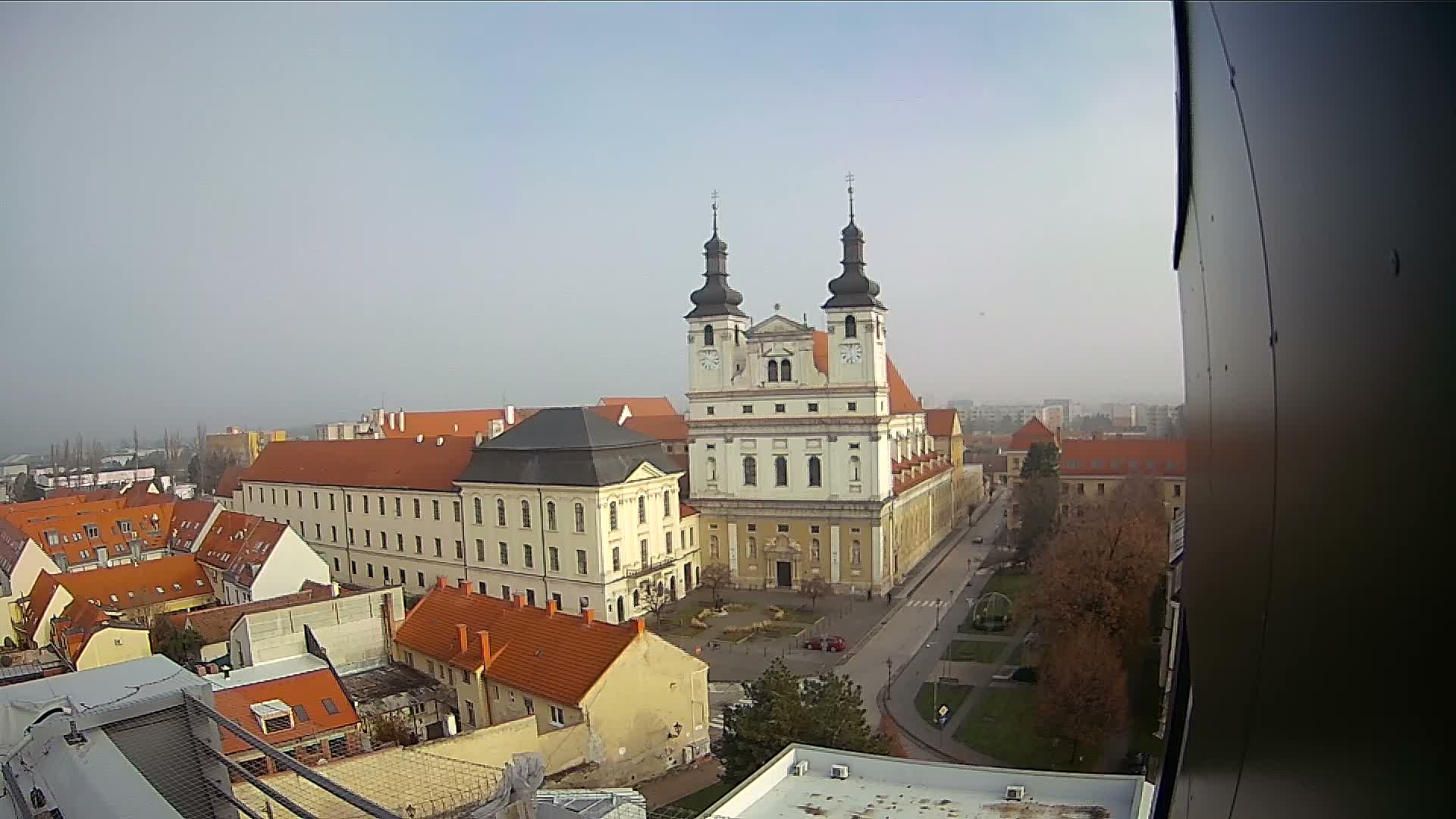 An aerial view showcases a European town dominated by a large, ornate white church with twin spires, surrounded by buildings with reddish-orange roofs, all beneath a uniformly hazy, overcast sky.