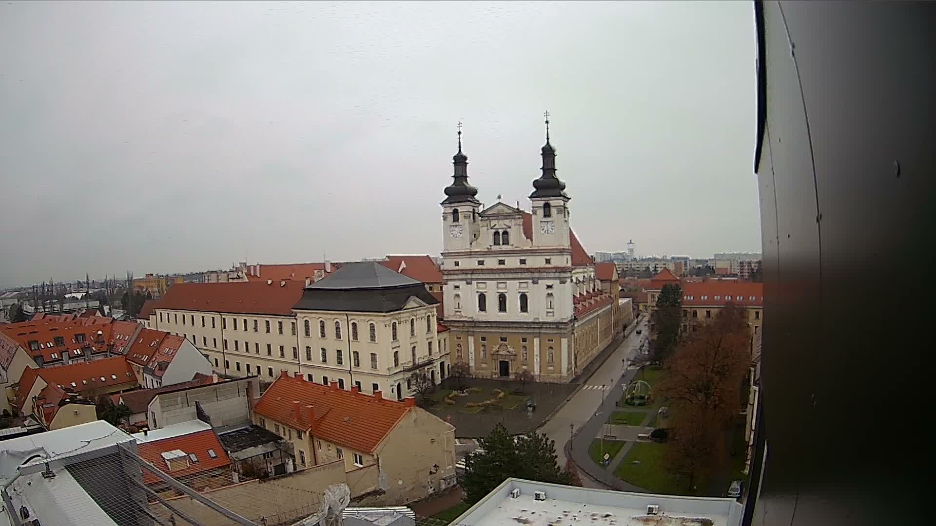 An elevated urban landscape reveals a grand baroque church with two dark-domed towers and white facades, surrounded by historic buildings with red-tiled roofs and a street, all under a uniformly overcast sky.