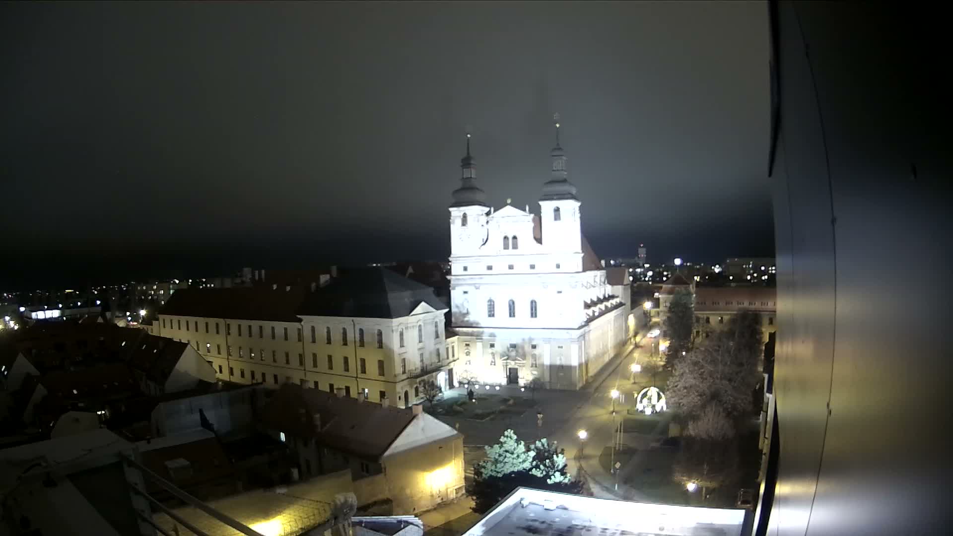 An elevated urban landscape reveals a grand baroque church with two dark-domed towers and white facades, surrounded by historic buildings with red-tiled roofs and a street, all under a uniformly overcast sky.
