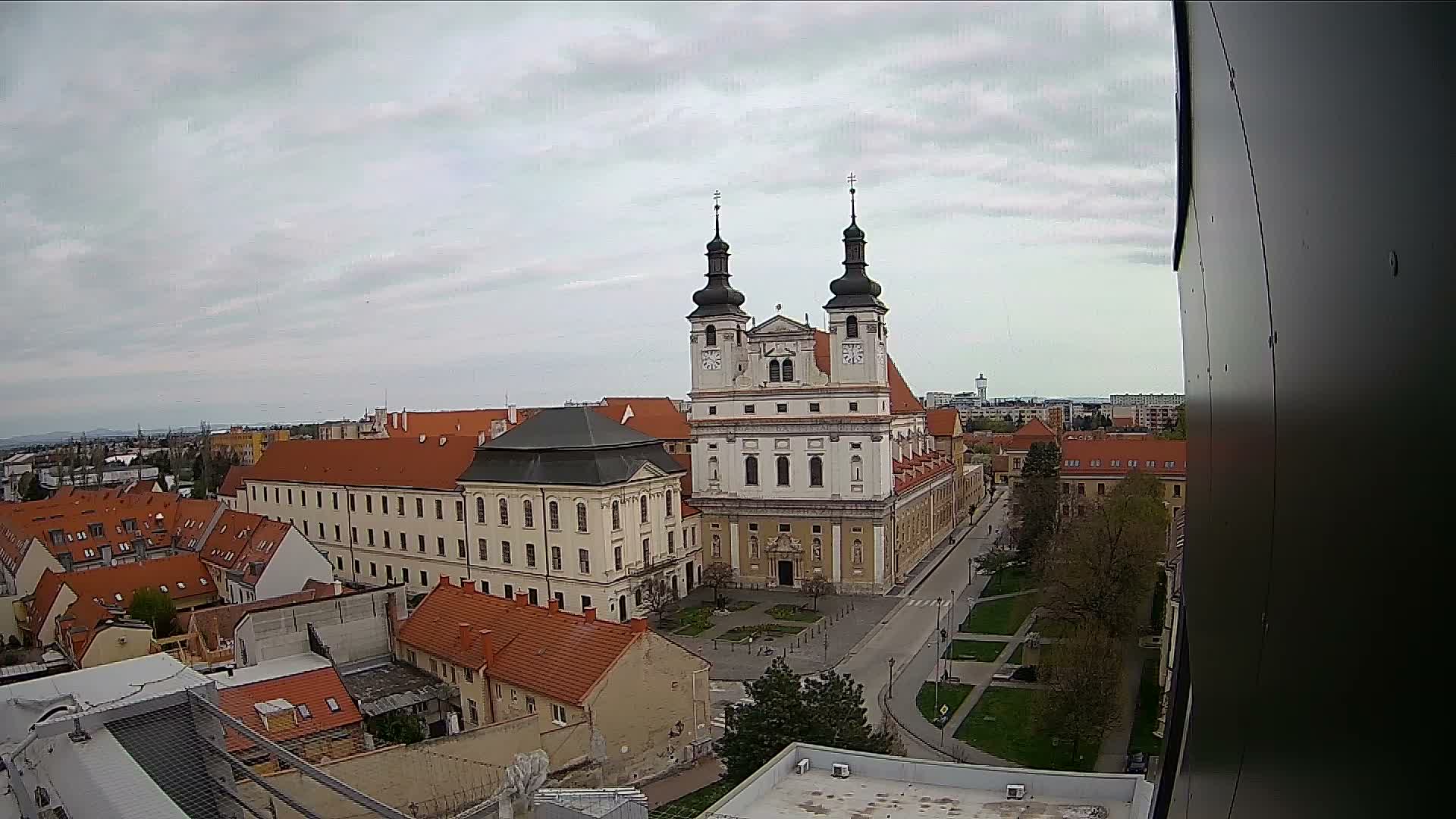 An elevated urban landscape reveals a grand baroque church with two dark-domed towers and white facades, surrounded by historic buildings with red-tiled roofs and a street, all under a uniformly overcast sky.