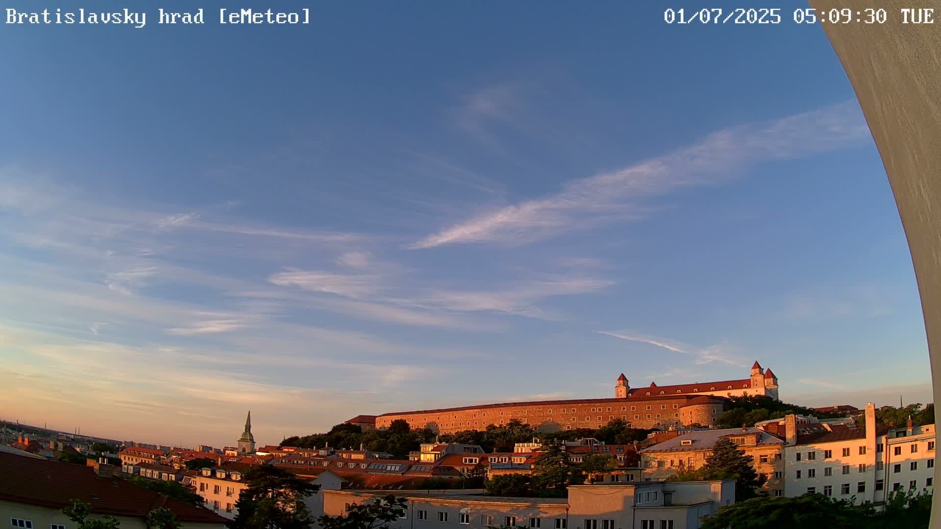 A large stone castle sits atop a hill overlooking a city under a mostly clear, light blue sky with wispy clouds during sunrise.