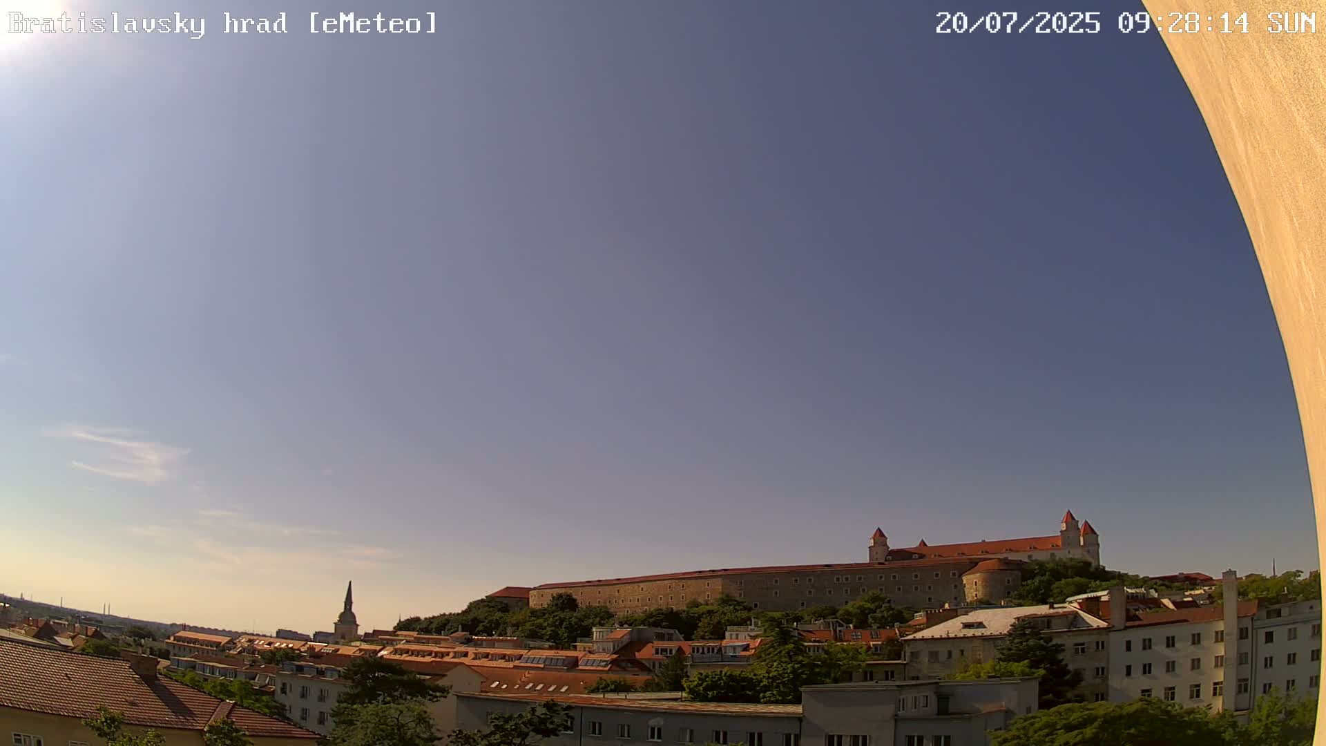 A large castle sits atop a hill overlooking a city of low-rise buildings under a clear, light-blue sky.