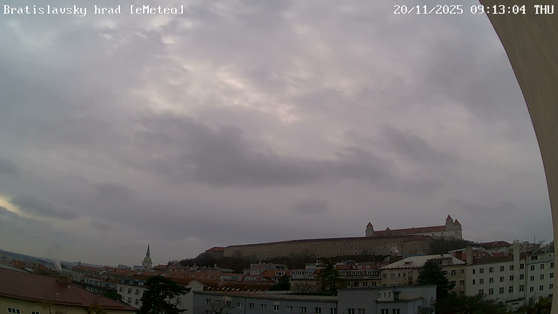 A panoramic view shows Bratislava Castle perched on a hill above a cityscape featuring numerous buildings and a distinct church spire, all under a uniformly gray and overcast sky.
