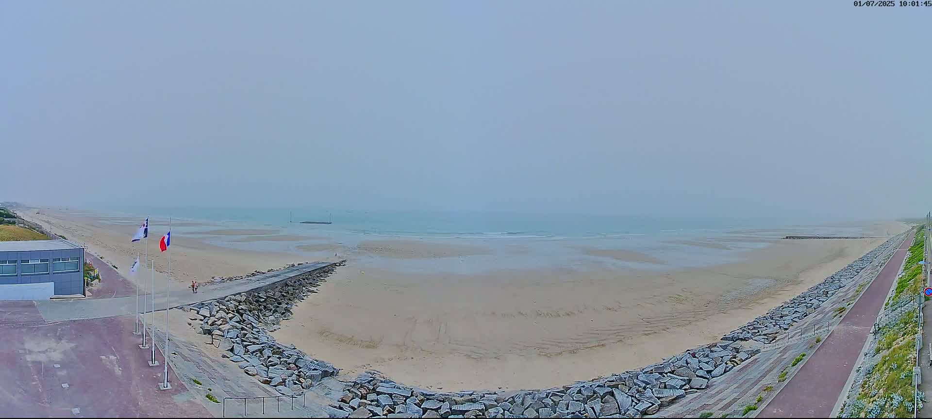 A foggy day reveals a sandy beach bordered by a rocky path and a building, with flags waving near a walkway leading to the water.