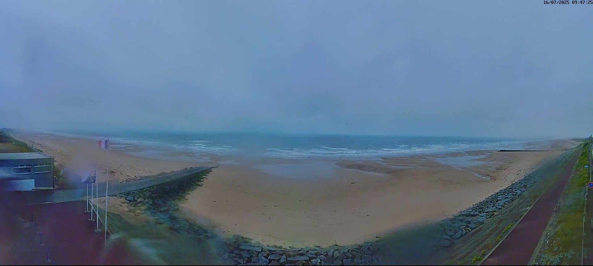A misty, overcast day reveals a wide sandy beach with a calm ocean, bordered by a paved path and a low stone wall.