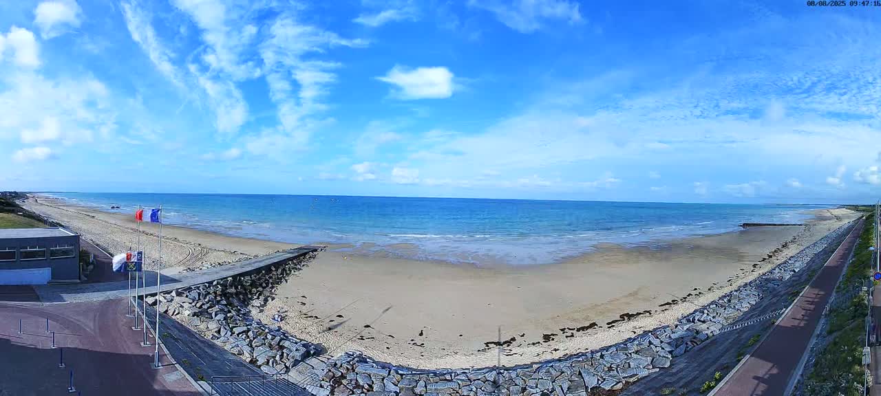 A sandy beach with calm ocean waves under a mostly sunny sky is seen from a high vantage point, showing a rocky seawall, a paved pathway, and a building near the beach.