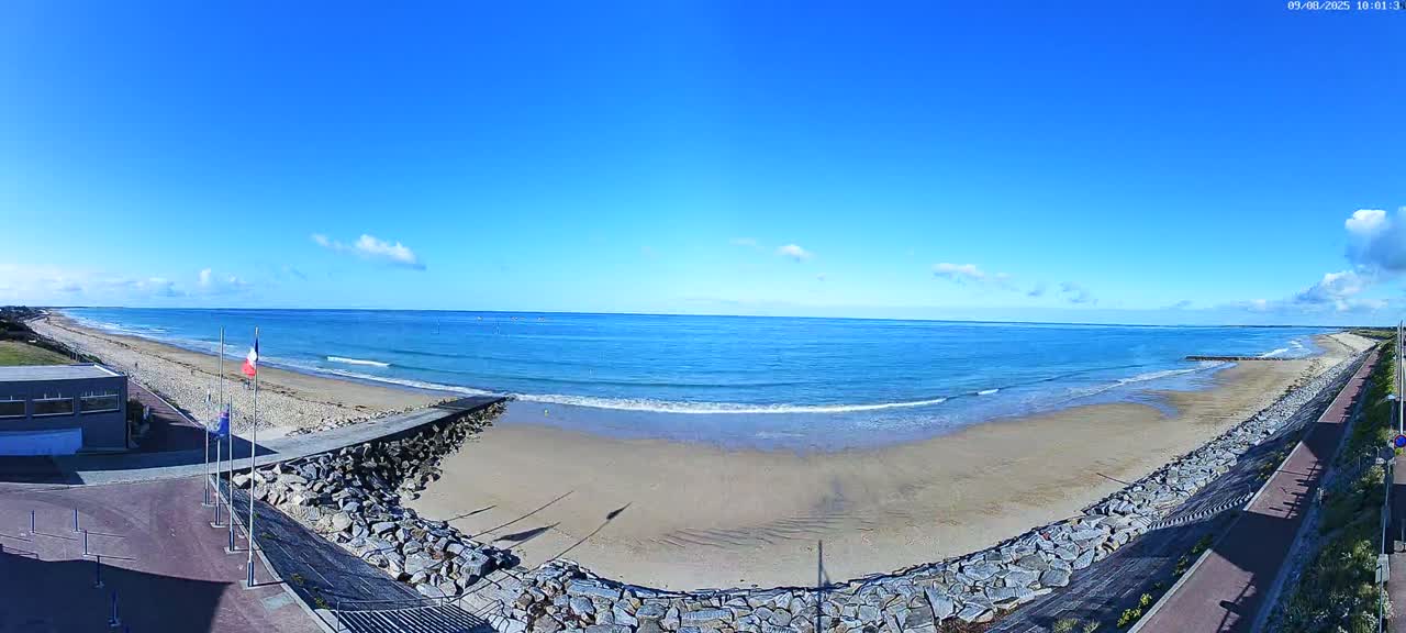A sandy beach with small waves stretches along a calm, blue ocean under a mostly clear, sunny sky.