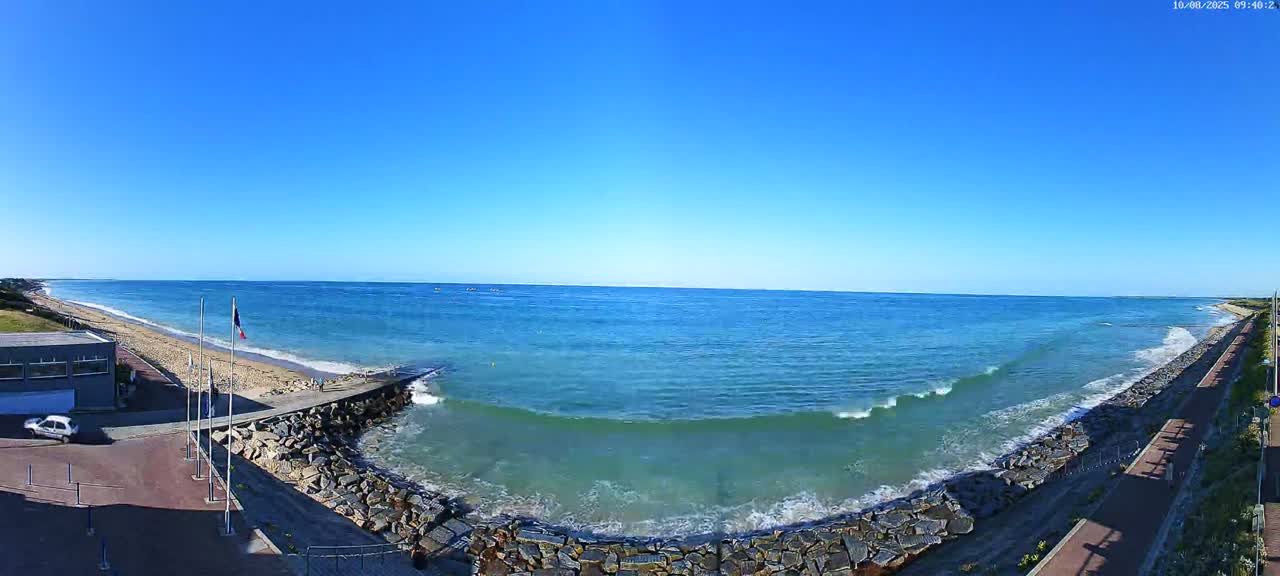 A panoramic view of a beach on a sunny day shows a calm ocean with small waves breaking against a rocky shoreline, a sandy beach, and a paved walkway.