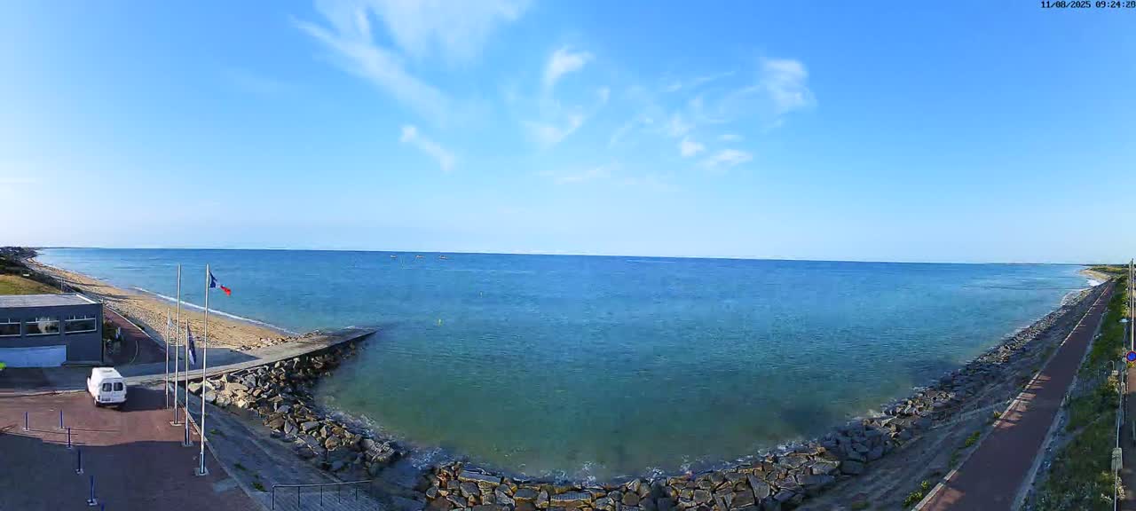 A mostly sunny day shows a wide expanse of calm blue ocean meeting a sandy beach backed by a paved path and a small building.