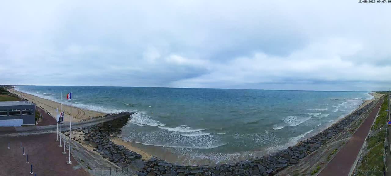 A wide, panoramic view shows a partly cloudy day at a sandy beach with a rocky shoreline and a building near a flagpole, with small waves breaking on the shore.