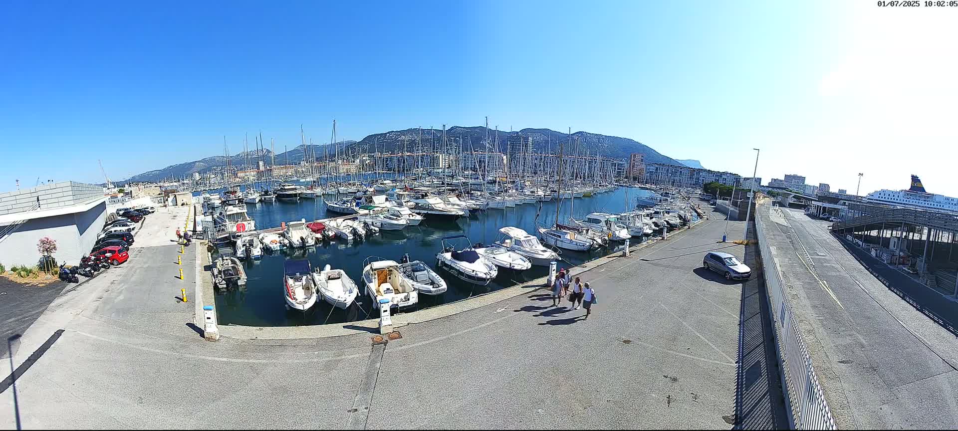 A marina filled with numerous boats and yachts is situated near a city under a clear, sunny sky.