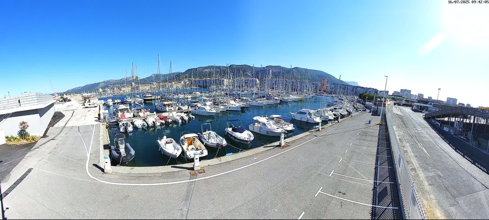 A marina filled with numerous boats of various sizes is situated near a city with mountains in the background under a clear, sunny sky.