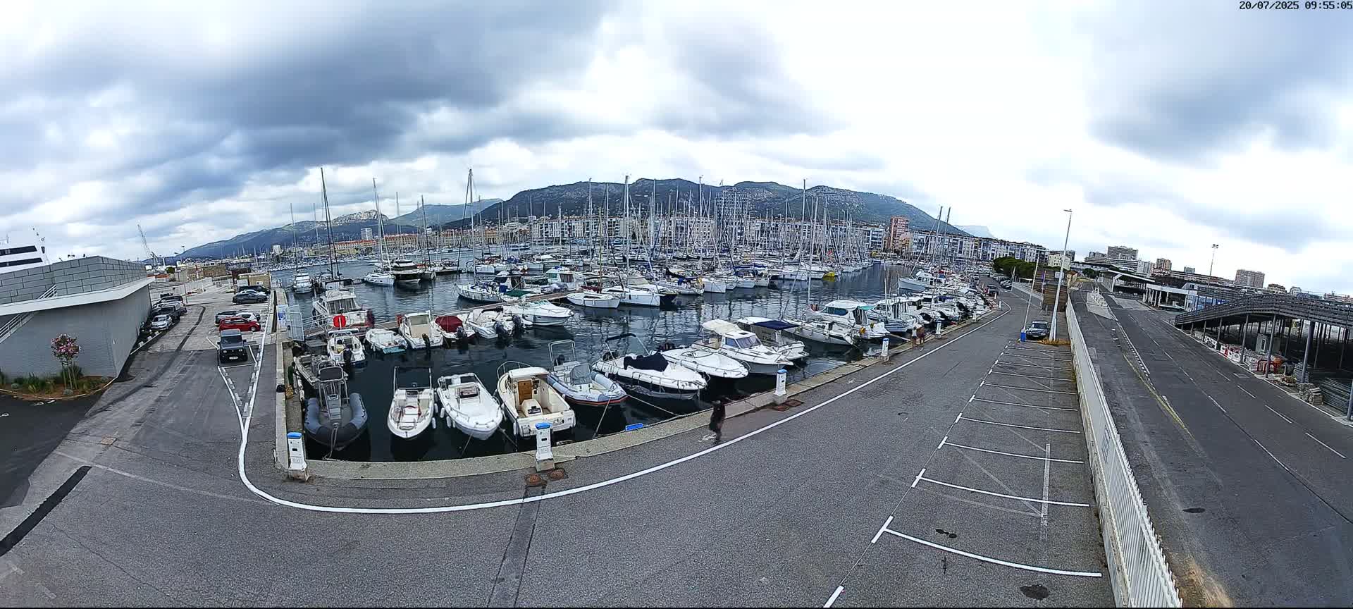 A marina filled with numerous boats is situated near a city under a cloudy sky, with a road and parking lot adjacent to the waterfront.